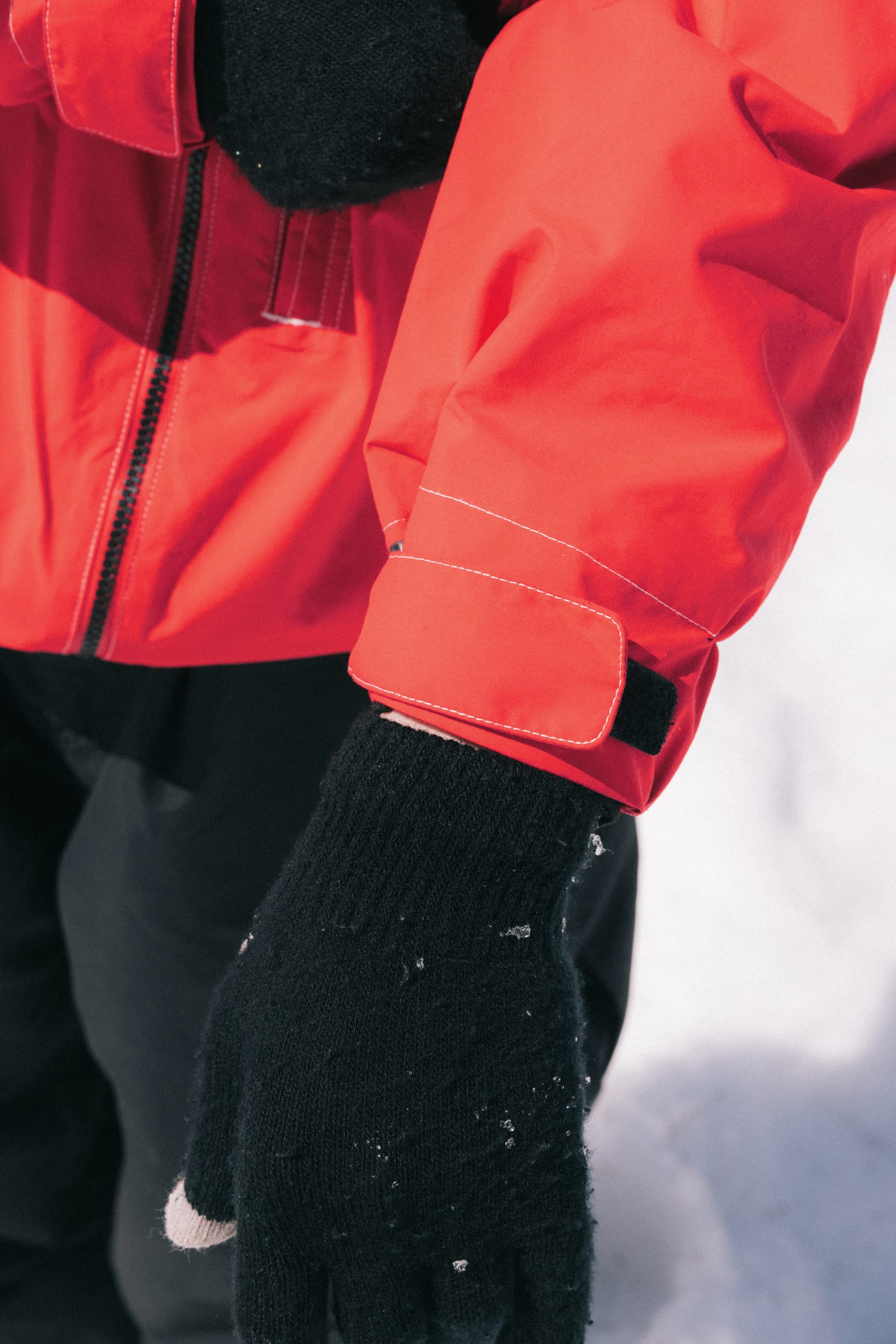 A person wearing the 686 Dojo® GORE-TEX Jacket in bright red by 686 and black gloves stands outdoors in the snow, with only their arm, part of their torso, and gloved hand visible.