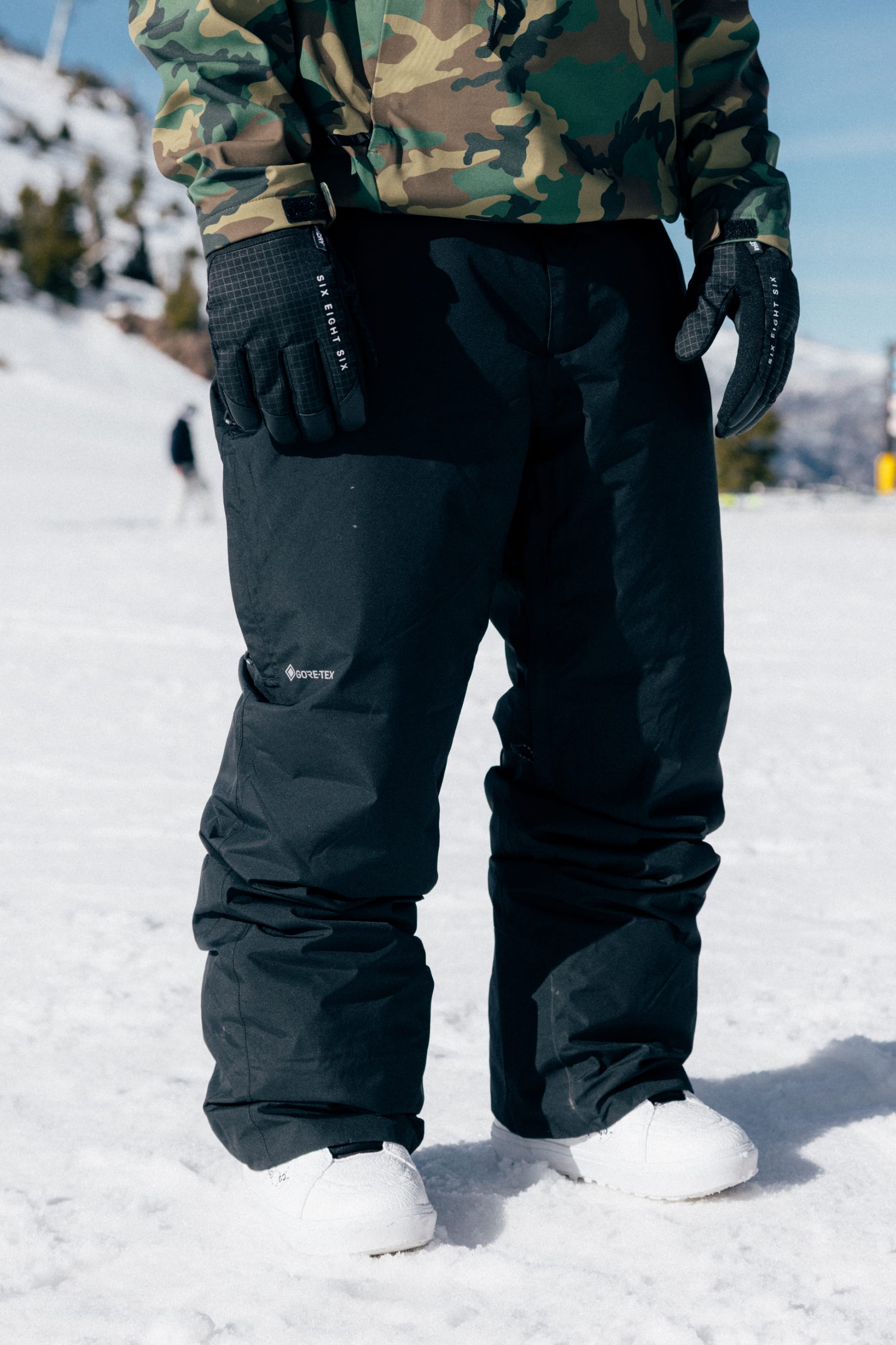 A person wearing 686 Men’s GORE-TEX® Core Shell Pant stands on snowy ground with trees and mountains in the background.