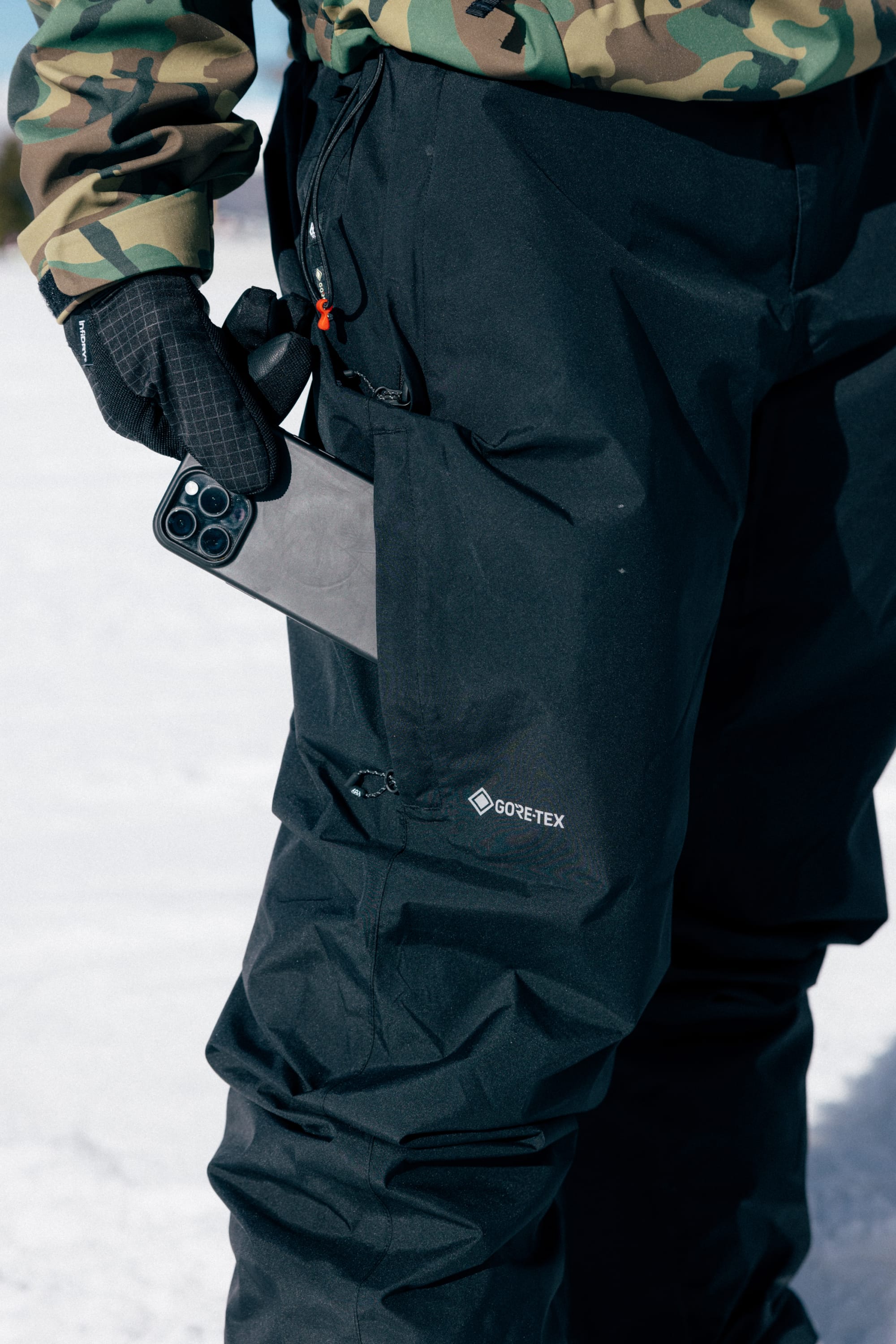 A person wearing 686 Men’s GORE-TEX® Core Shell Pants in black and a camouflage jacket stands on snow, holding a smartphone partially tucked into their pocket.