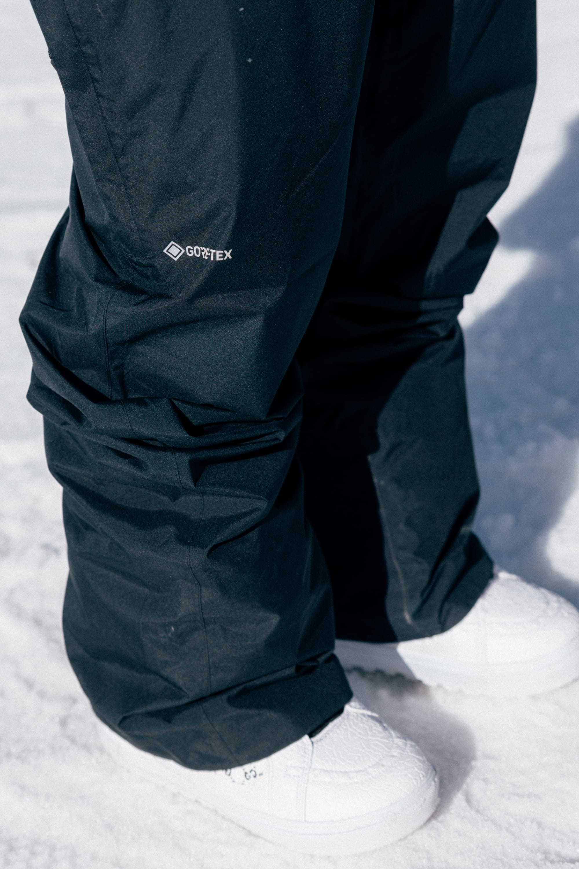 A person wearing 686 Men’s GORE-TEX® Core Shell Pants in black and white shoes stands on snow, with only the lower half of their legs visible.
