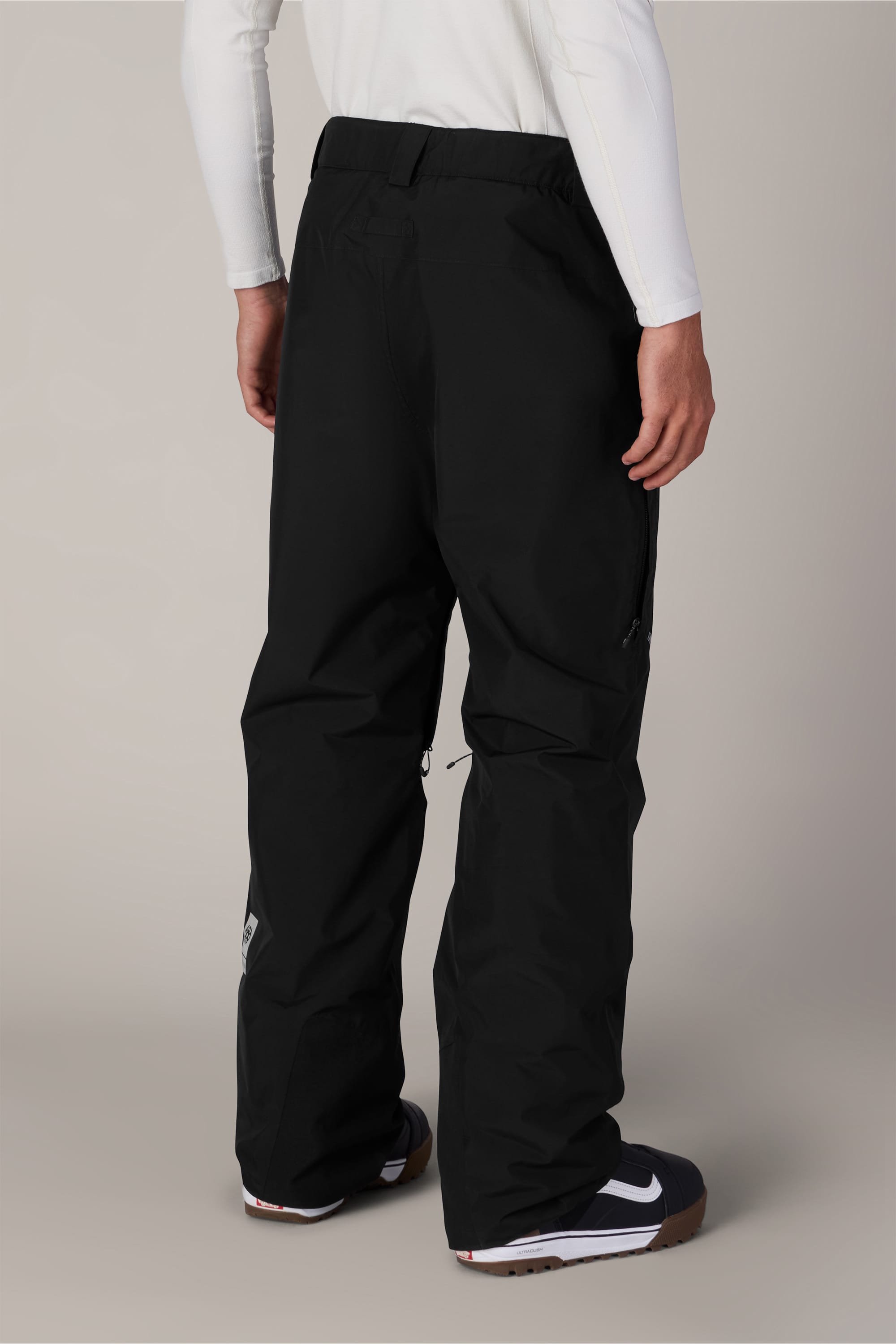 A person wearing 686 Men’s GORE-TEX® Core Shell Pant in black, a white long-sleeve shirt, and black Nike sneakers is standing facing away from the camera against a neutral background.