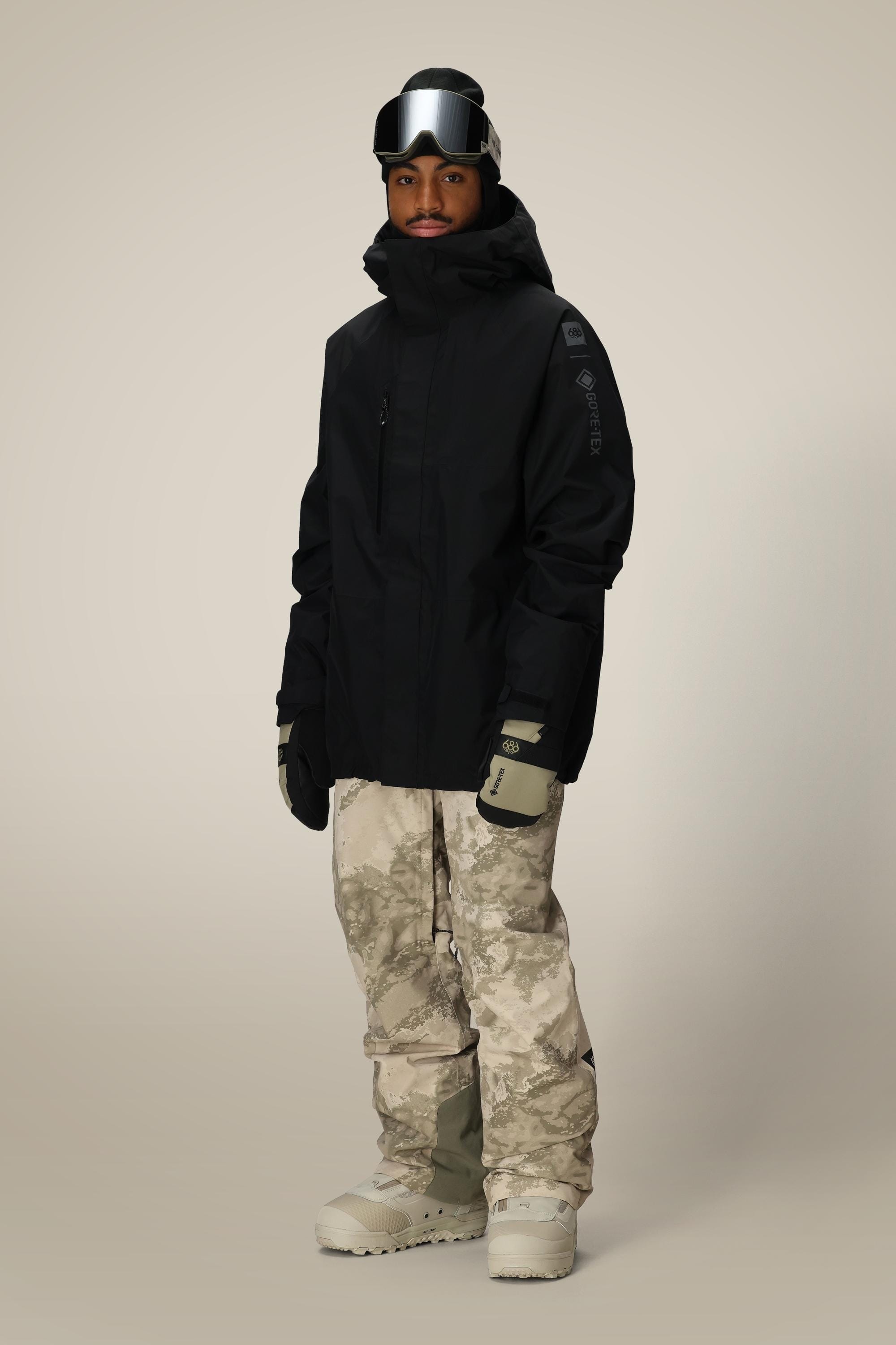 A person stands against a plain background wearing a black hooded jacket, 686 Mens GORE-TEX Core Shell Pant in tan and gray camo, light gray gloves, beige snow boots, and black ski goggles on their forehead.