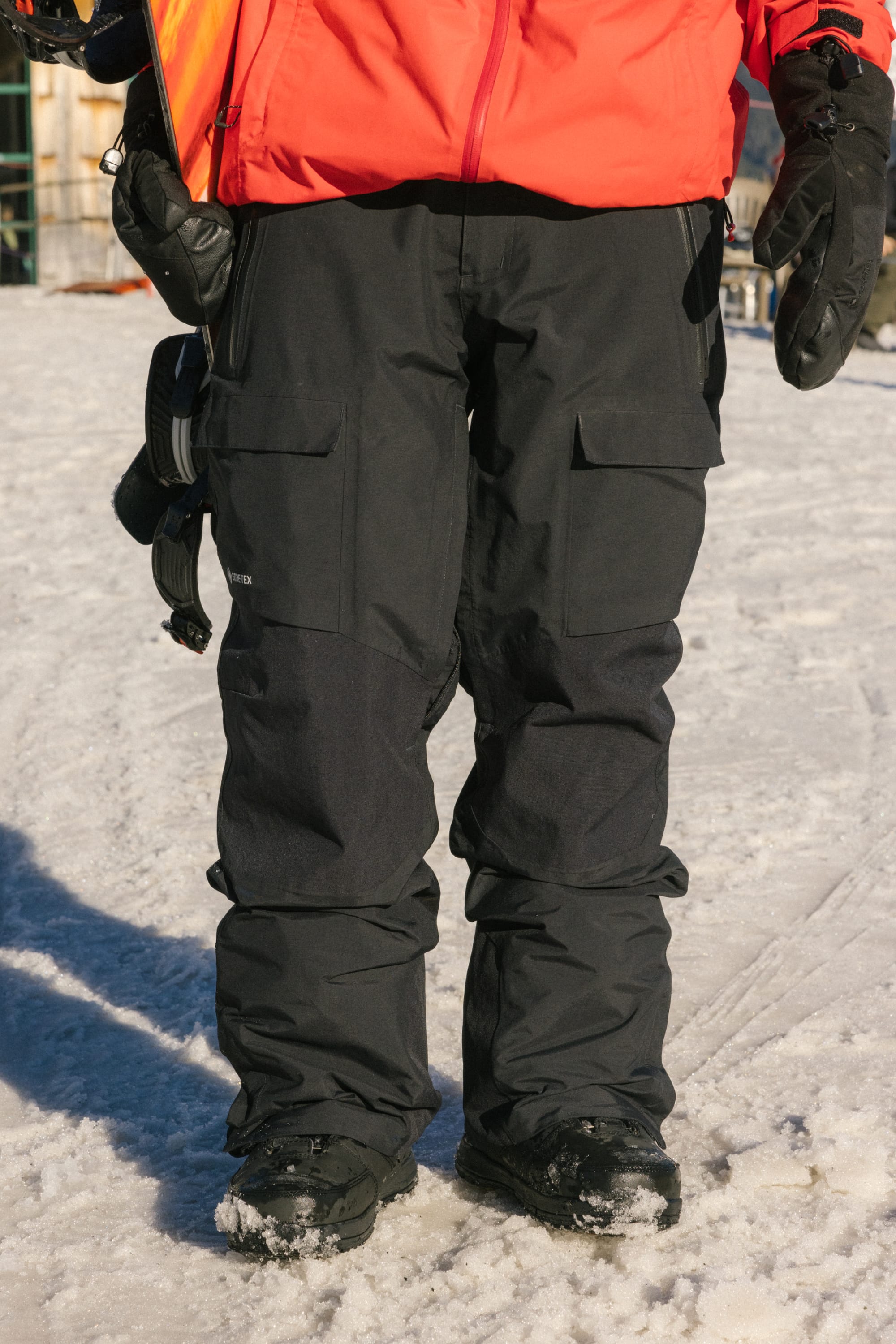 A person stands on snow wearing 686 Men's GORE-TEX Dispatch™ Shell Pant and a bright orange jacket, with black gloves and a snowboard binding visible by their side.