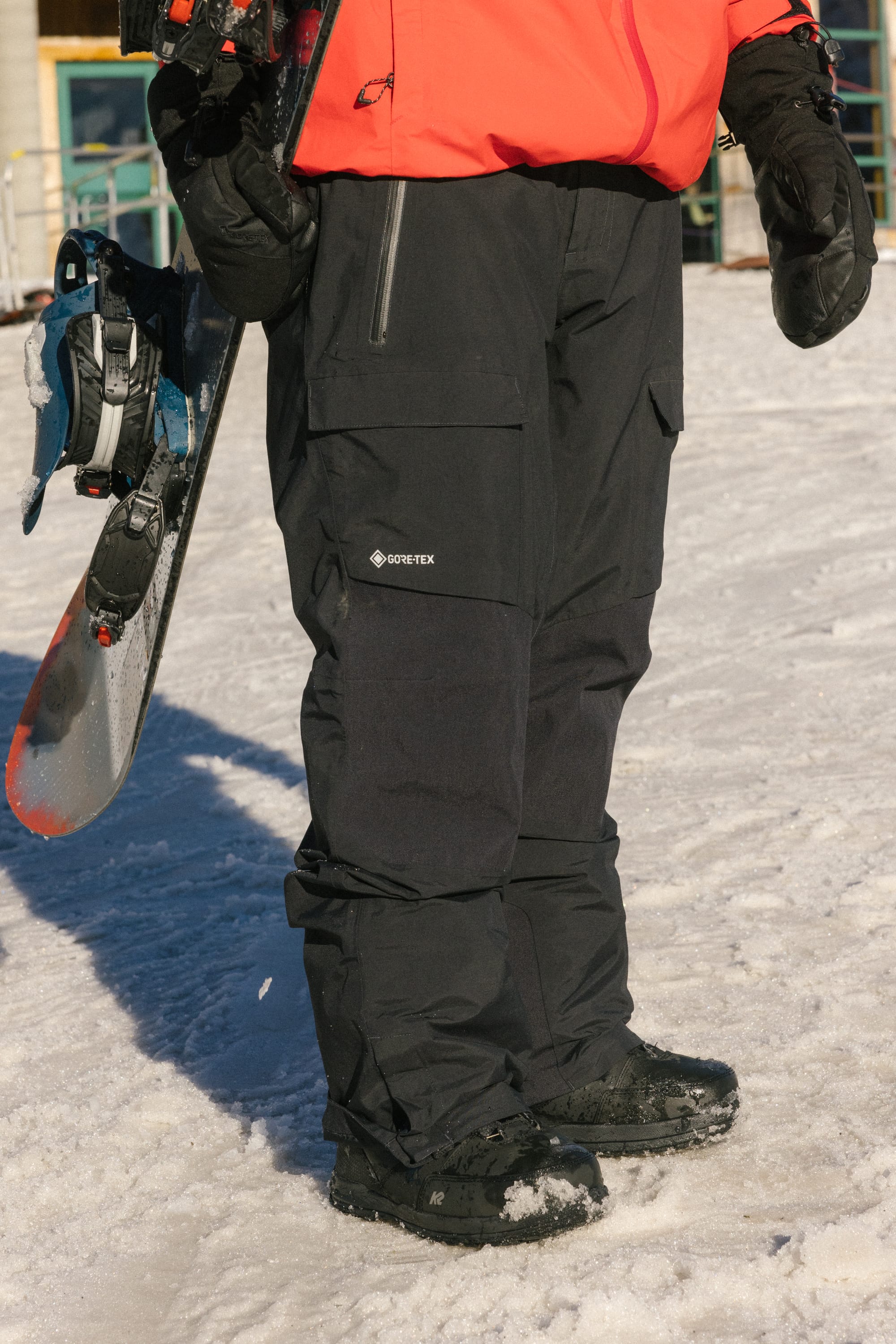 Wearing 686 Men's GORE-TEX Dispatch™ Shell Pants, a person in snow boots and gloves stands on snow with a snowboard upright. Only their lower body is visible, along with a red jacket.