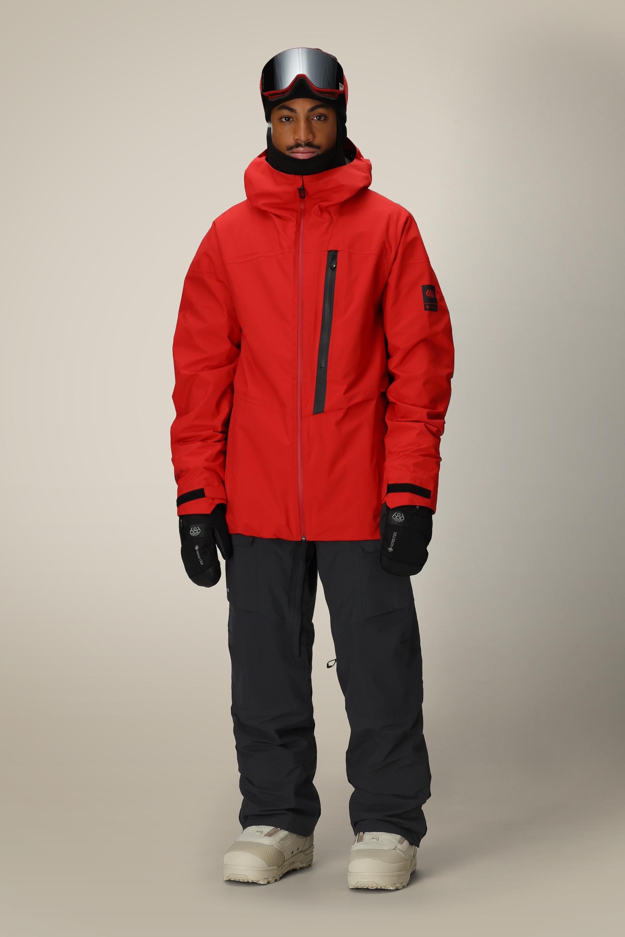 A person wearing a red ski jacket, 686 Men's GORE-TEX Dispatch™ Shell Pant by 686, white boots, black gloves, a black helmet, and ski goggles stands facing forward against a plain beige background.