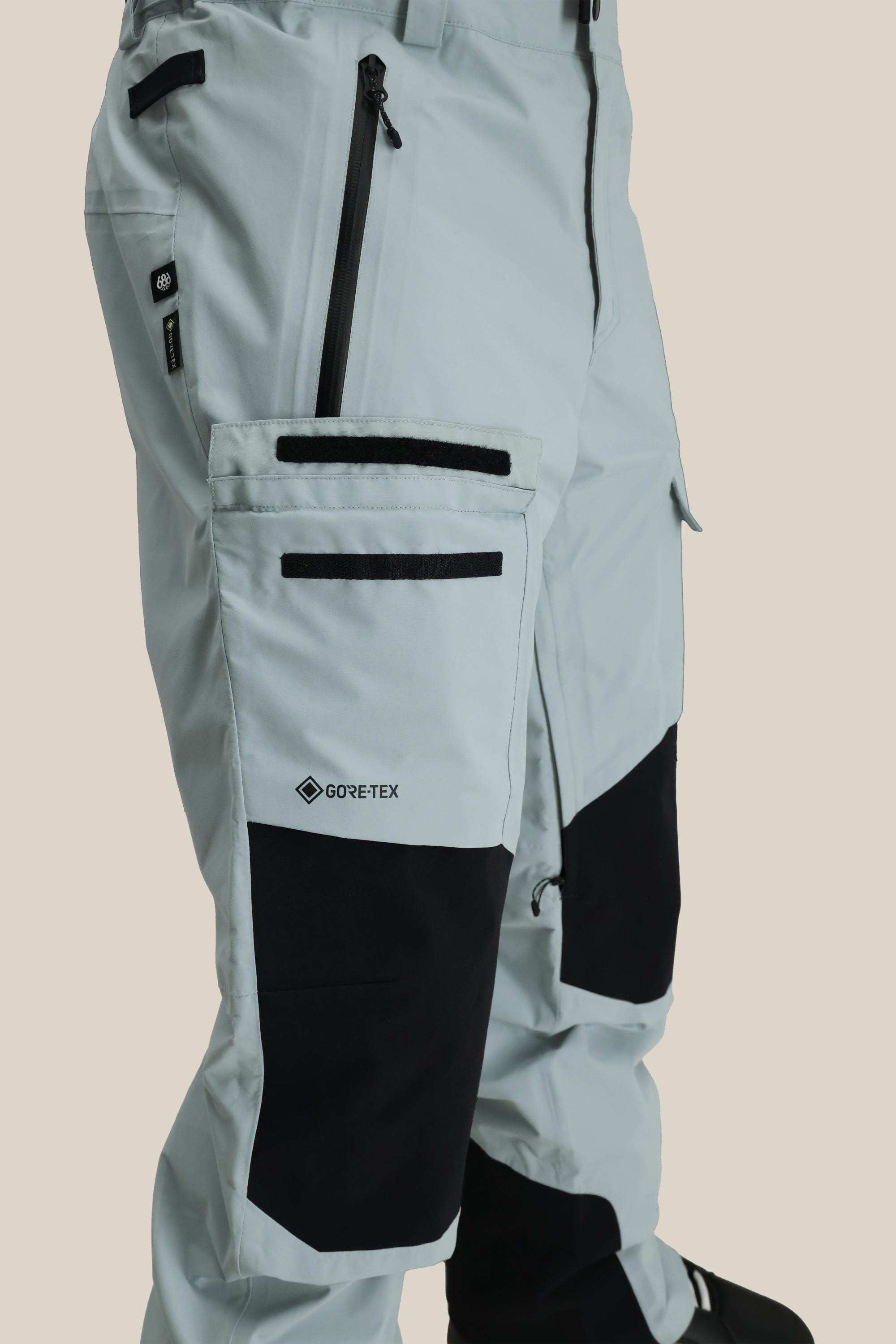 Close-up of the 686 Men's GORE-TEX Dispatch™ Shell Pant in light gray with black knee patches, zippered pockets, and a GORE-TEX logo on the cargo pocket. These durable nylon pants by 686 are ideal for outdoor activities.