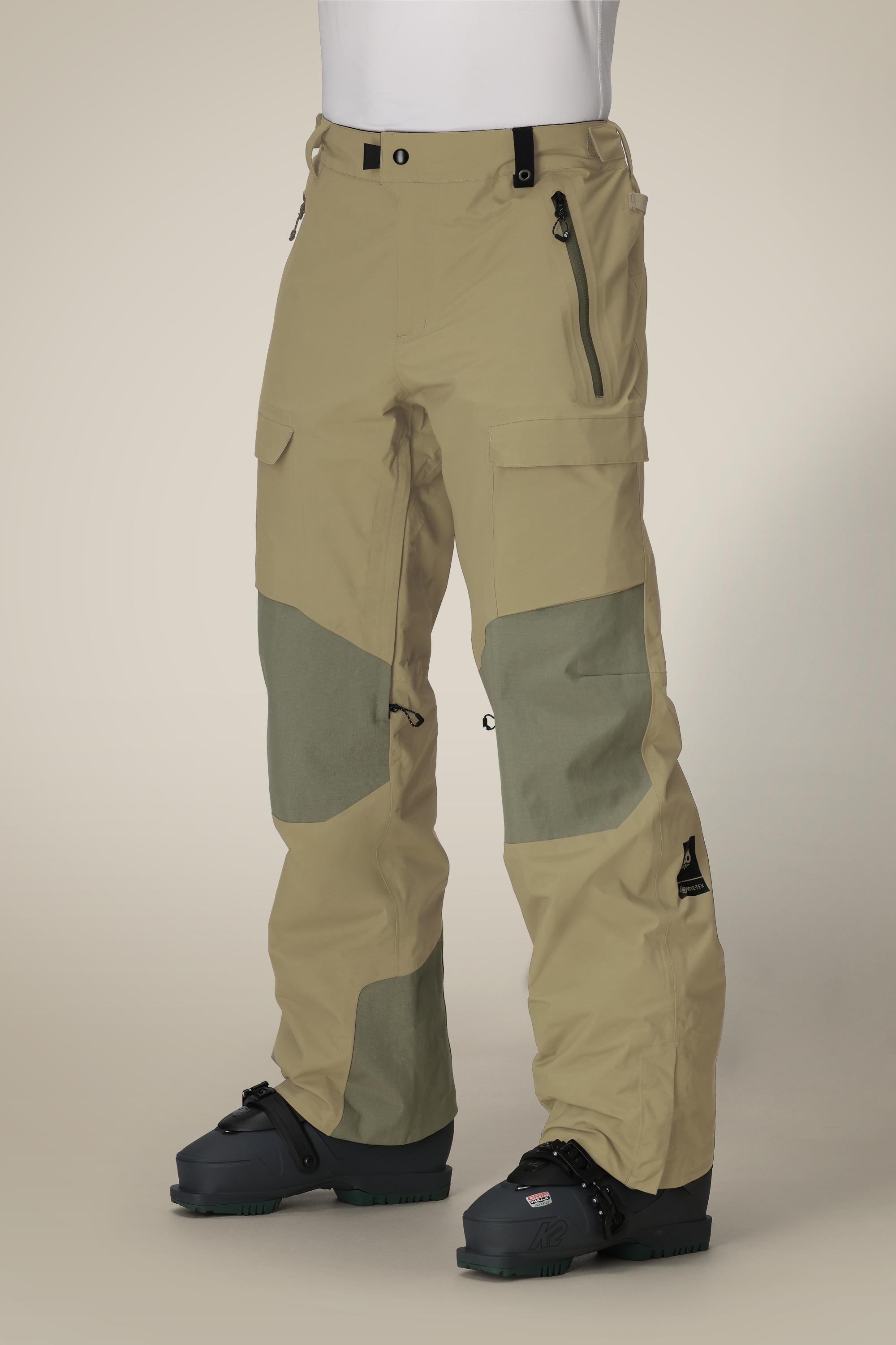 A person wears 686 Men's GORE-TEX Dispatch™ Shell Pant in beige and olive green, featuring zippered pockets and reinforced knee patches, paired with black snow boots, standing against a plain background.
