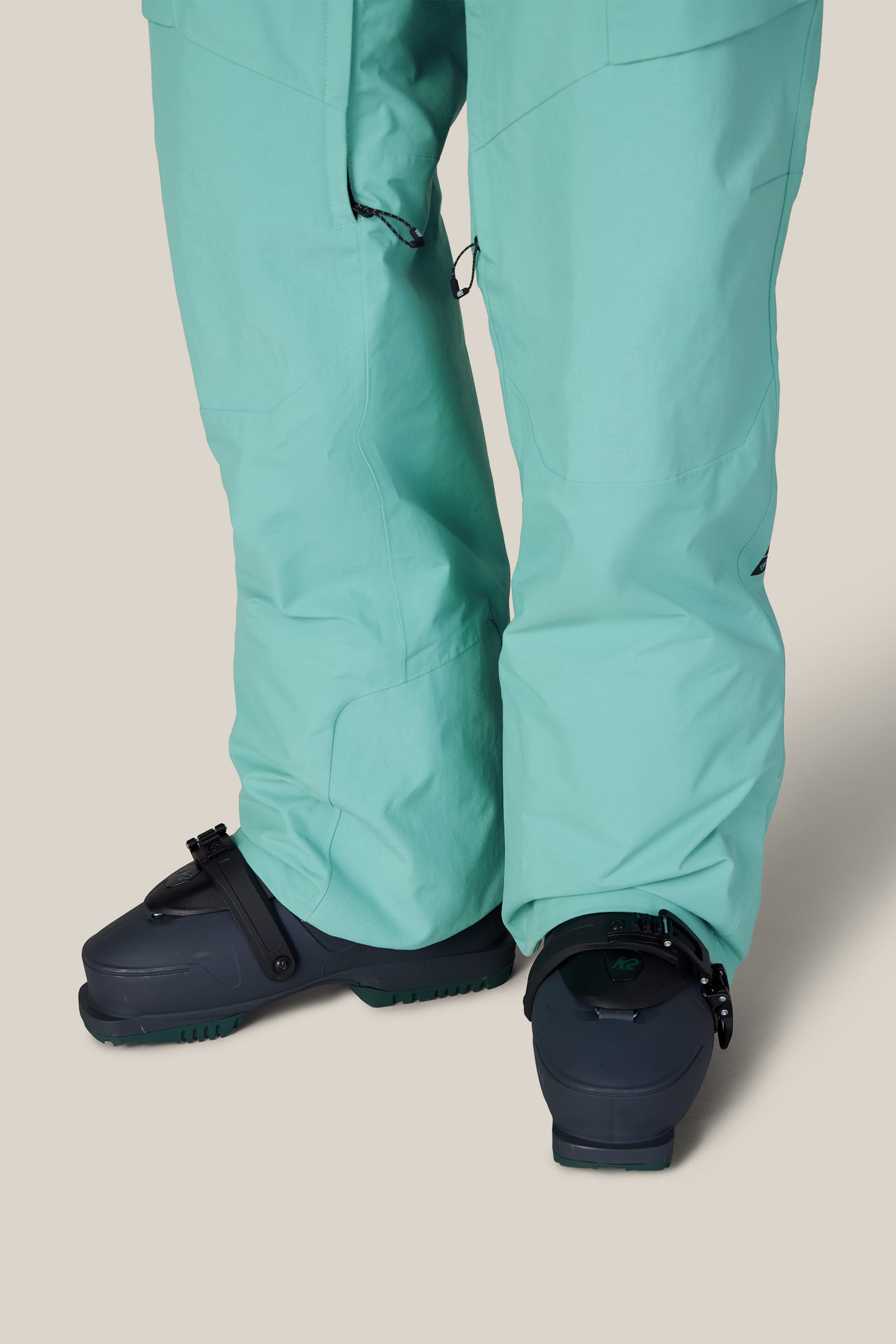 A person wearing 686 Men’s GORE-TEX® Dispatch™ Shell Pant in light teal and dark gray snowboard boots stands on a beige background. The slightly baggy snow pants from 686 cover part of the boots.
