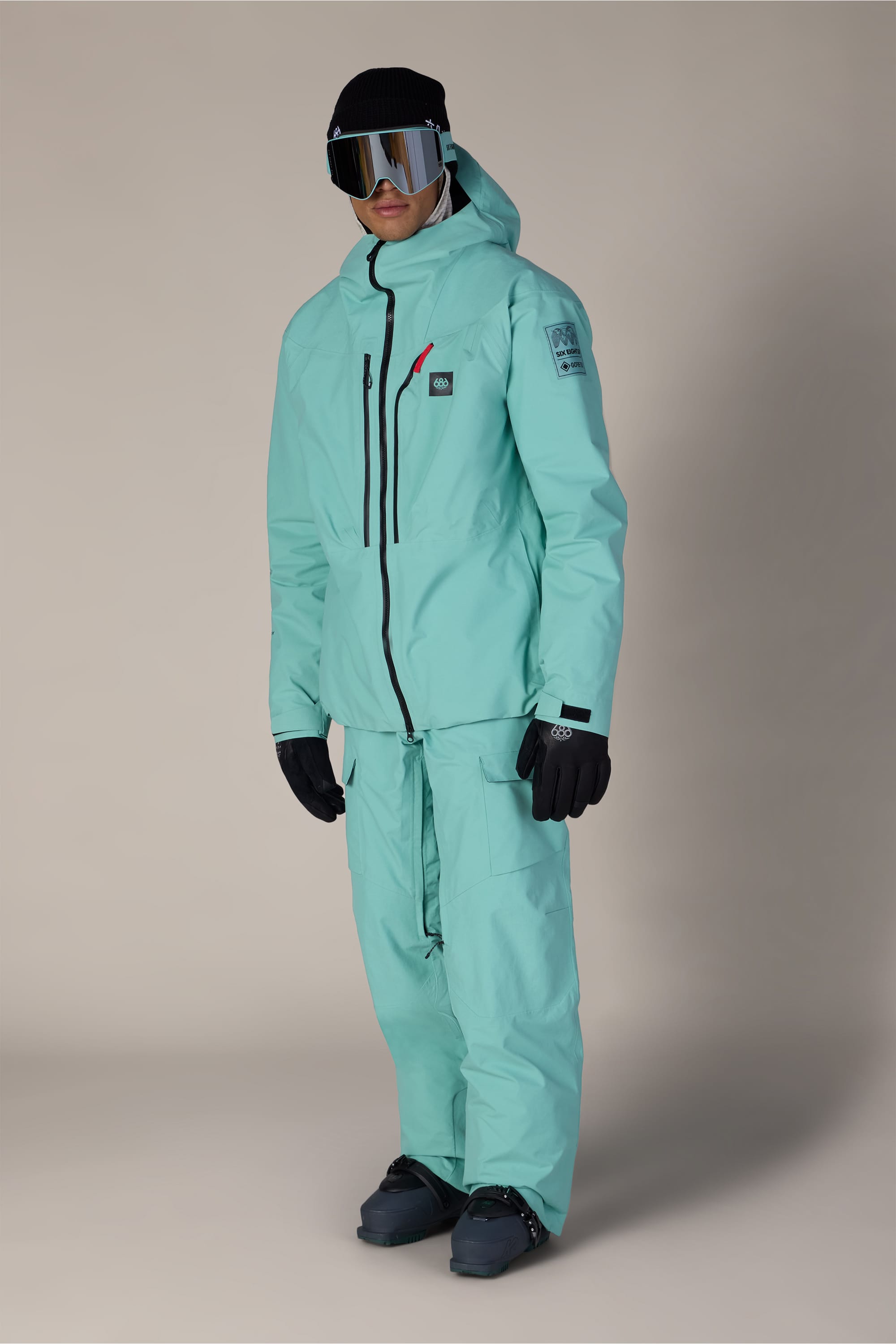 A person wearing a light teal snow jacket and the 686 Men’s GORE-TEX® Dispatch™ Shell Pant, paired with black gloves, black snow boots, ski goggles, and a black beanie, stands against a plain light background.
