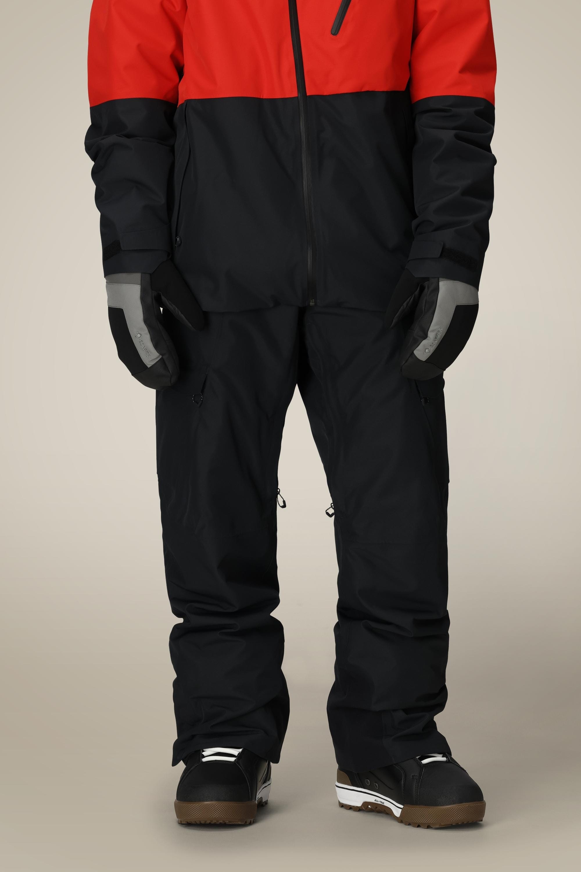 A person wearing a red and black winter jacket, black gloves, 686 Men's Quantum Thermagraph® Pant by 686, and black snow boots stands against a plain background. Their face and upper body are not visible.