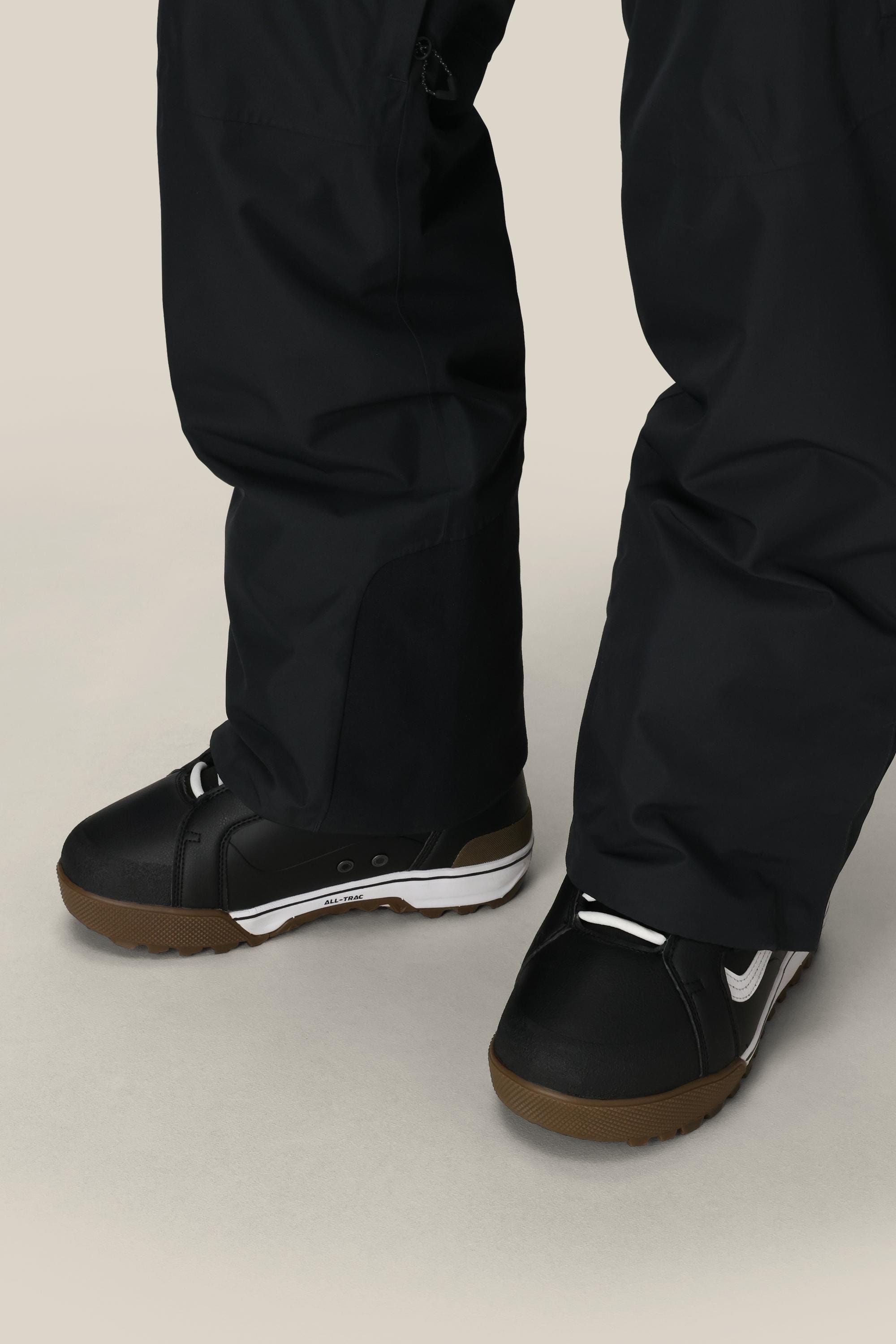 A close-up of a person wearing 686 Men's Quantum Thermagraph® Pant and black snowboard boots with brown soles, standing on a light-colored surface.