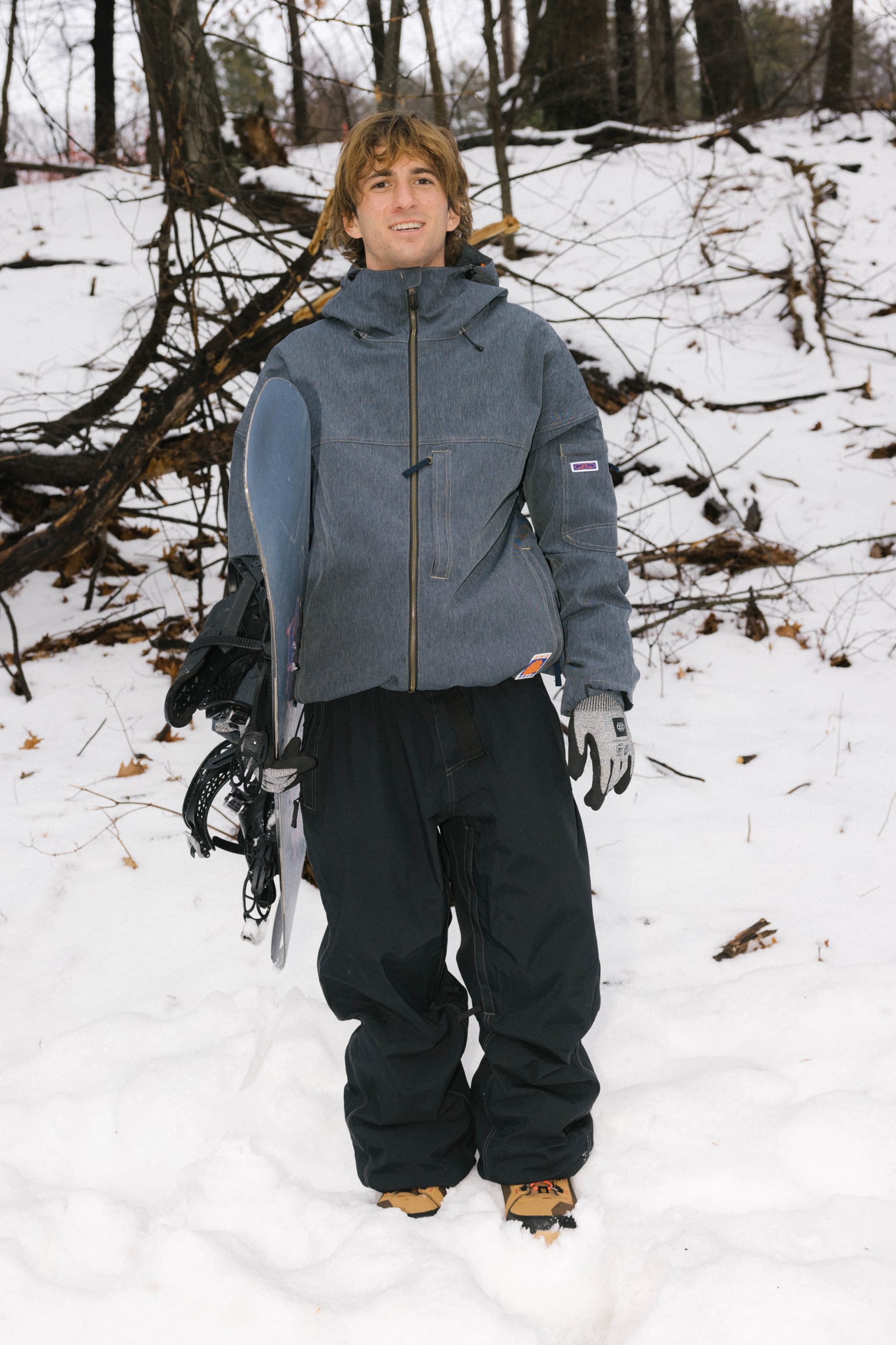 A person with long hair stands on snow, wearing a gray jacket, 686 Dojo® Pant by 686, gloves, and boots, holding a snowboard. Bare trees and branches fill the background.