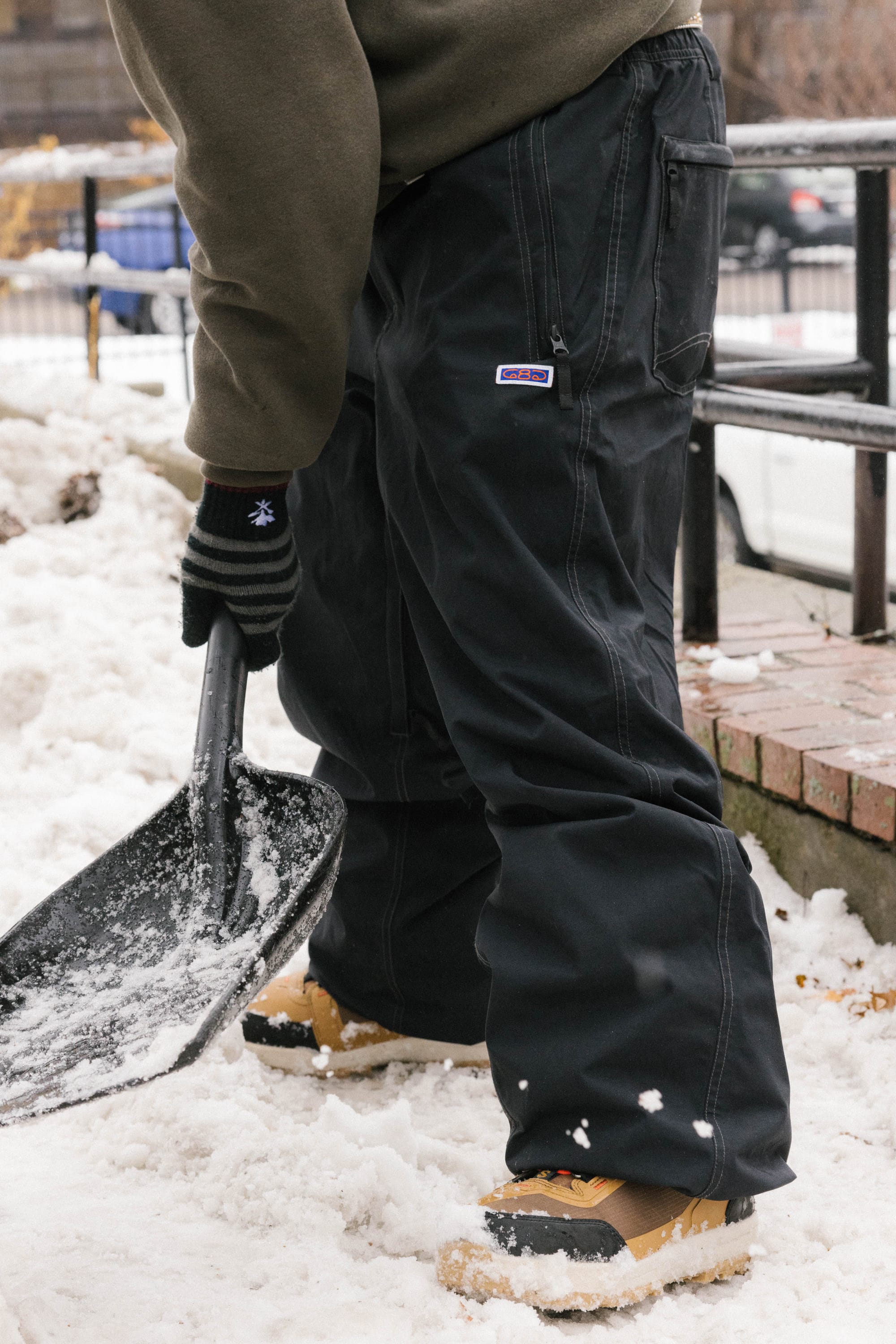 A person wearing black gloves, tan boots, and 686 Dojo® Pant by 686 shovels snow on a sidewalk. A brick wall, railing, and snow-covered ground appear in the background.