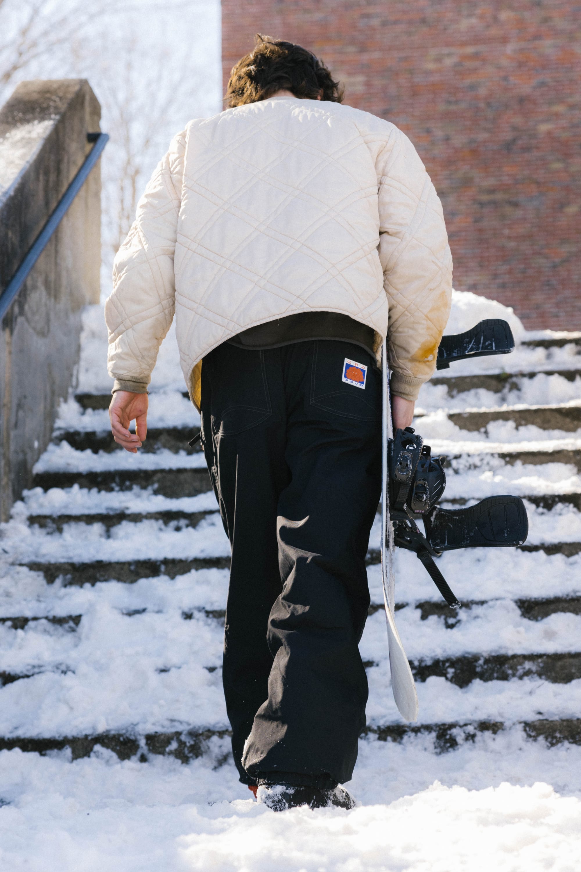 Wearing a cream quilted jacket and 686 Dojo® Pant by 686, a person walks up snowy steps holding a snowboard and camera in one hand.
