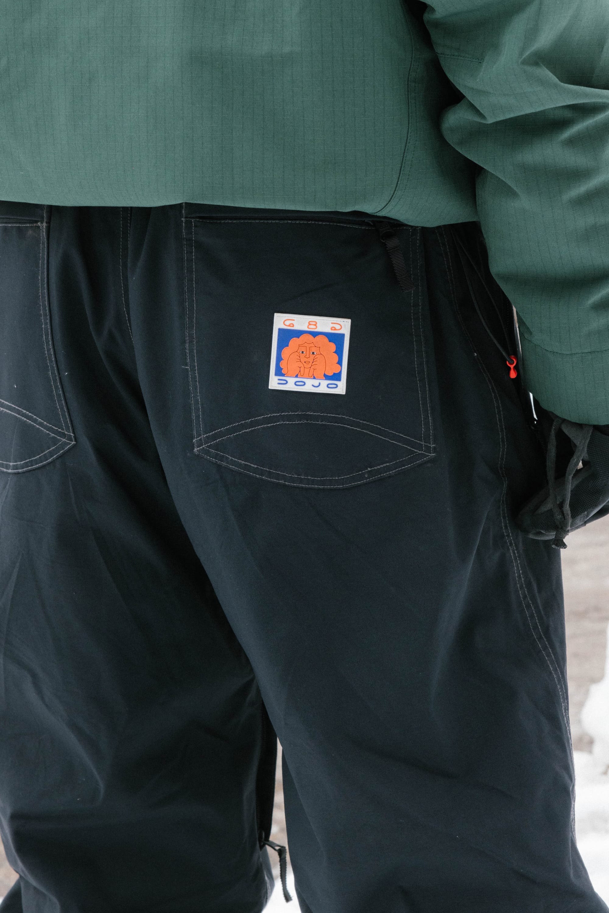 A person wearing the 686 Dojo® Pant by 686, featuring visible white stitching and a colorful "GAS CLOUD" patch on the back pocket, is seen with a green jacket in a snowy setting.