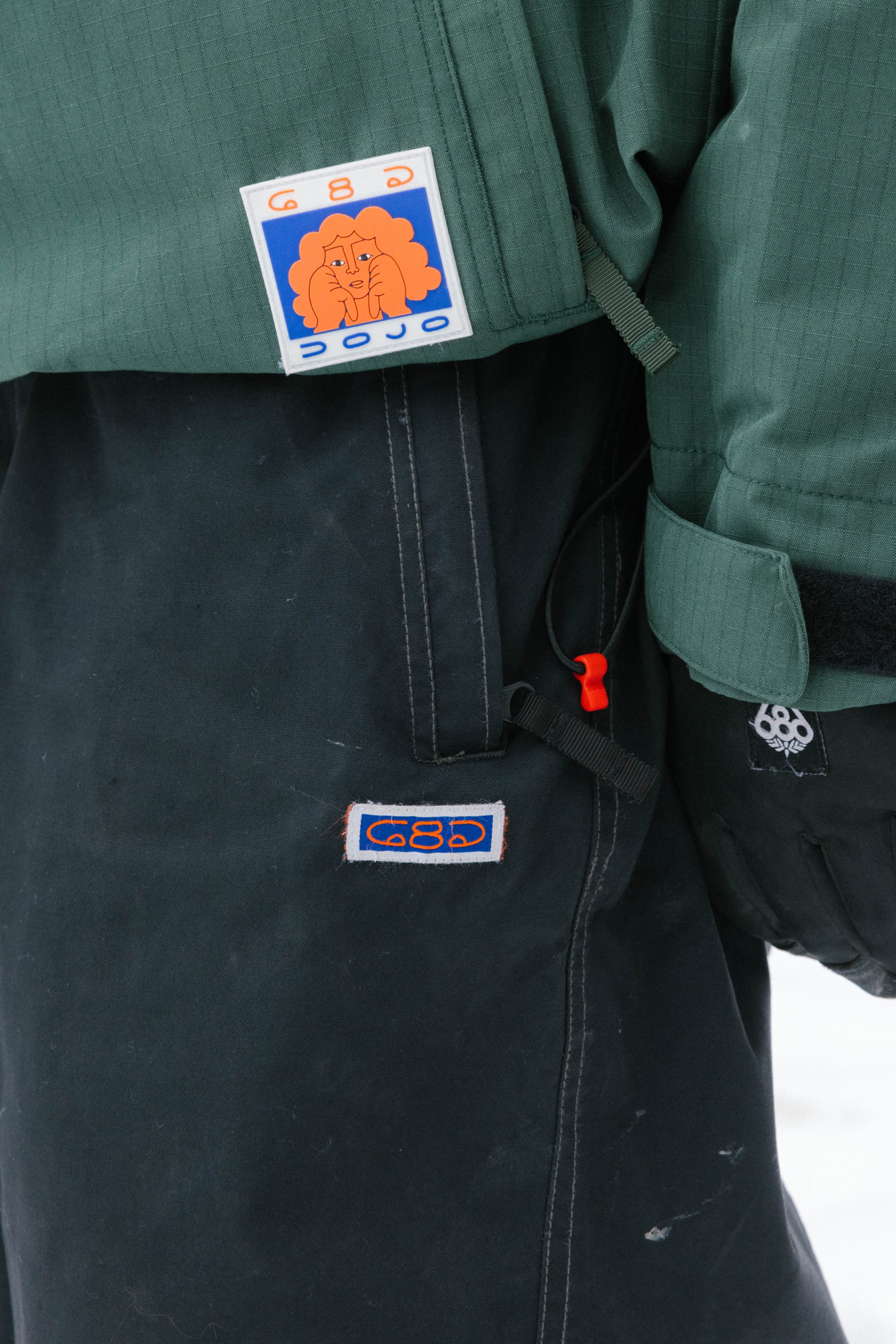 A close-up of a person wearing the 686 Dojo® Pant by 686—nylon waterproof pants in green and black, featuring colorful embroidered patches (one shows a cartoon face) and a red plastic toggle on the side.