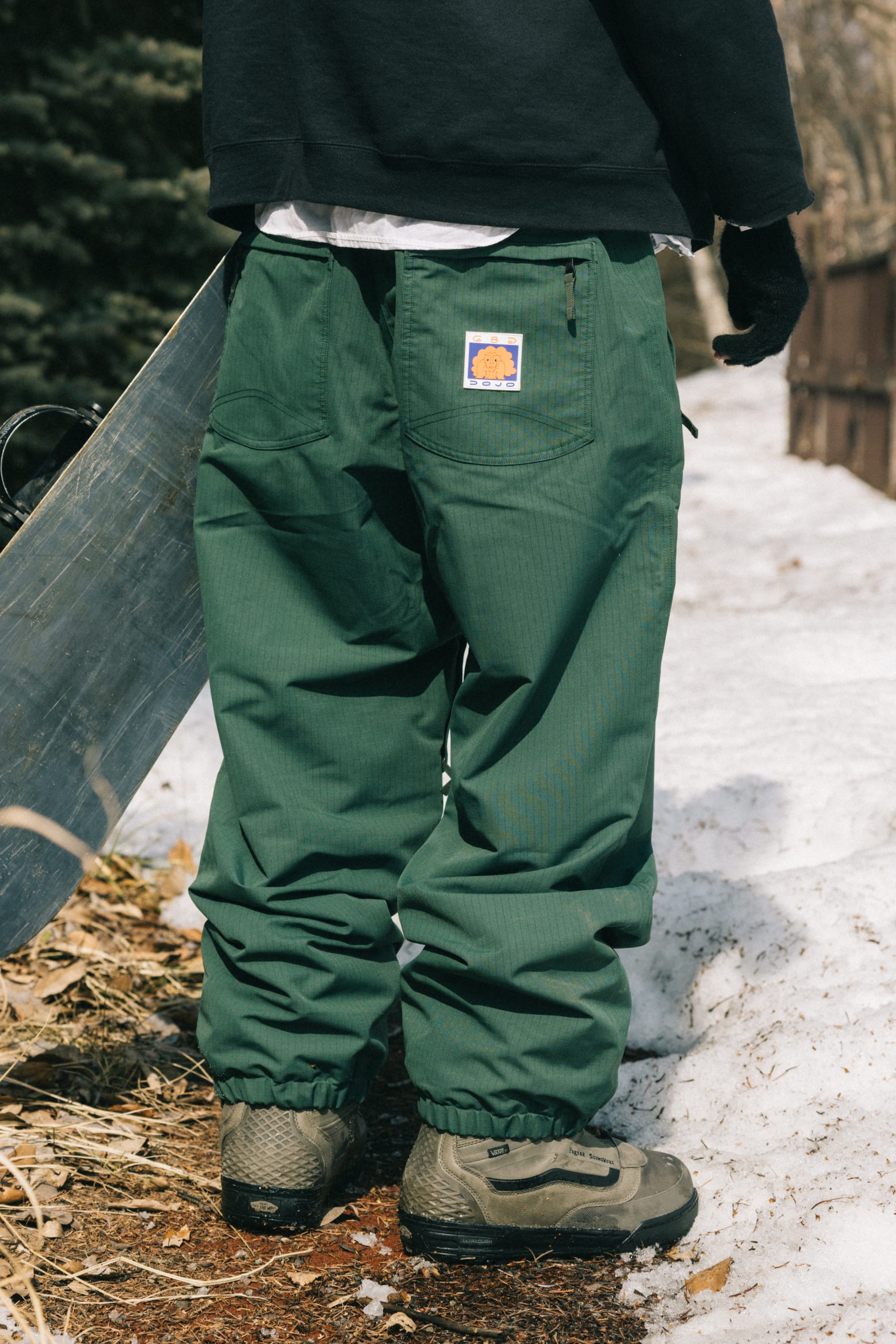 A person wearing 686 Dojo® Pant men's waterproof pants in green and gray boots stands on a snowy path with a snowboard. Pine trees and a wooden fence are in the background. The 686 pants feature a bear logo patch on the back pocket.