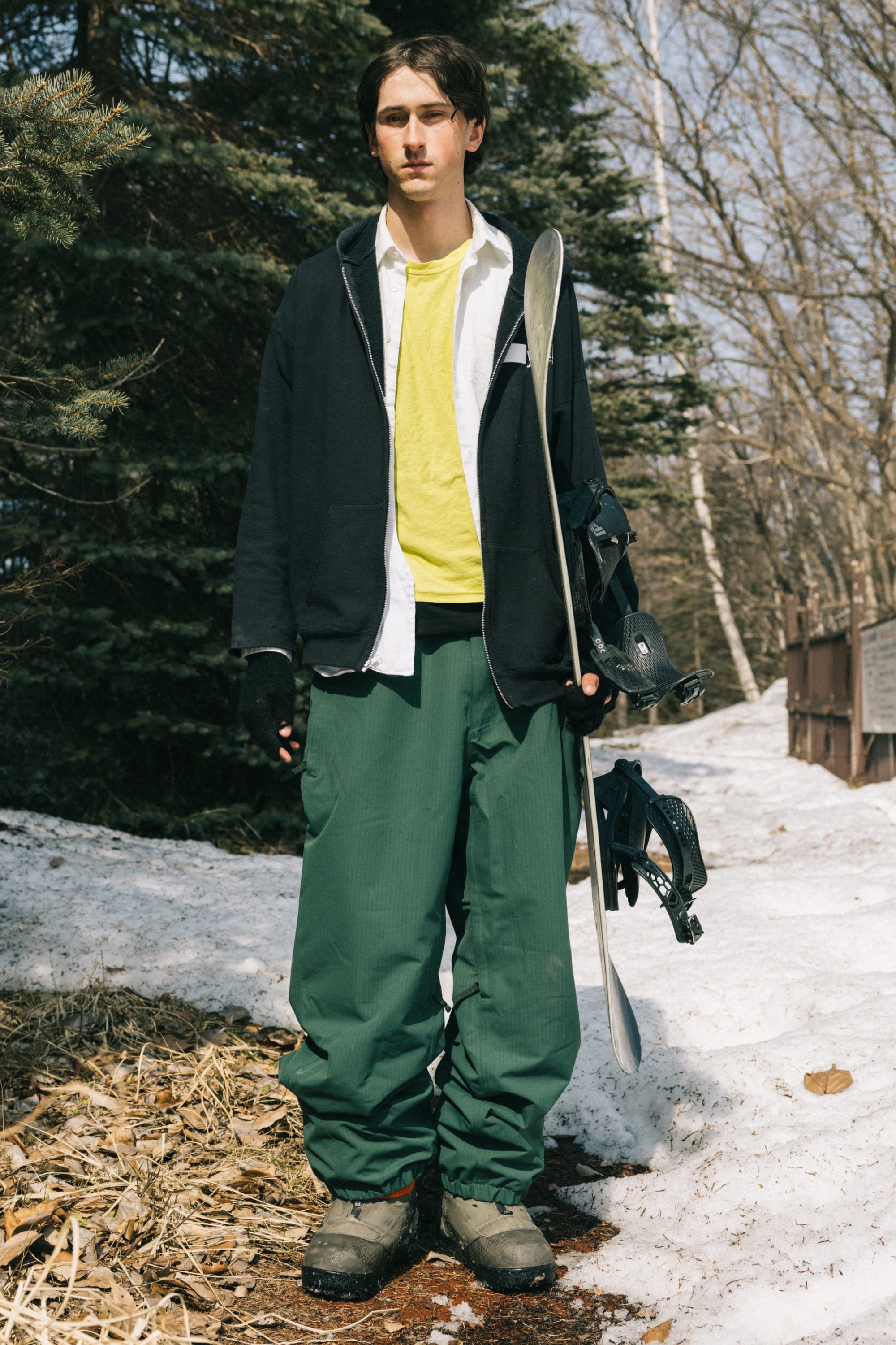 A young man stands outdoors on snow, wearing 686 Dojo® Pant by 686, a yellow shirt, a black zip-up jacket, and gloves while holding a snowboard. Trees and snowy patches appear in the background.