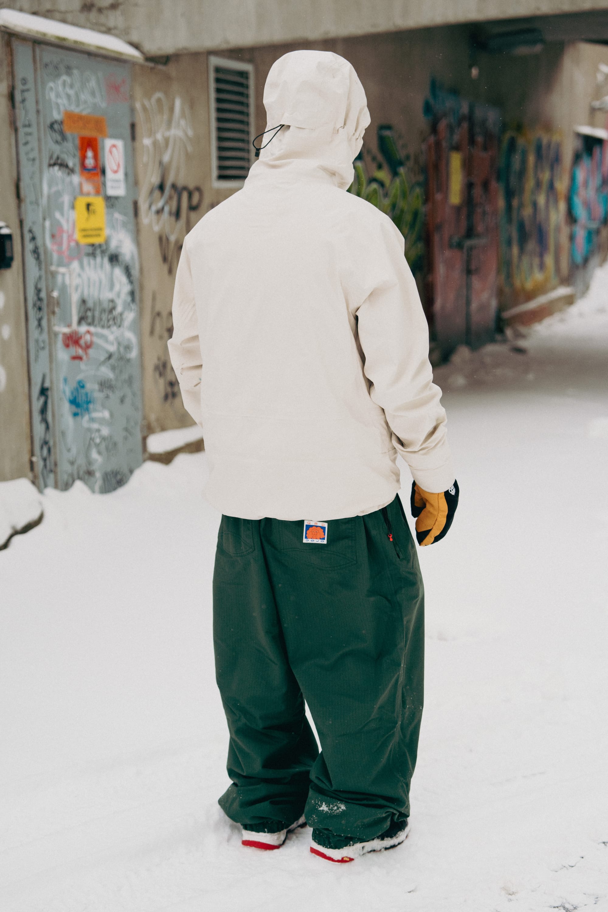 Wearing the 686 Dojo® Pant from 686, a person stands on snow in an alley with graffiti-covered walls, facing away from the camera.