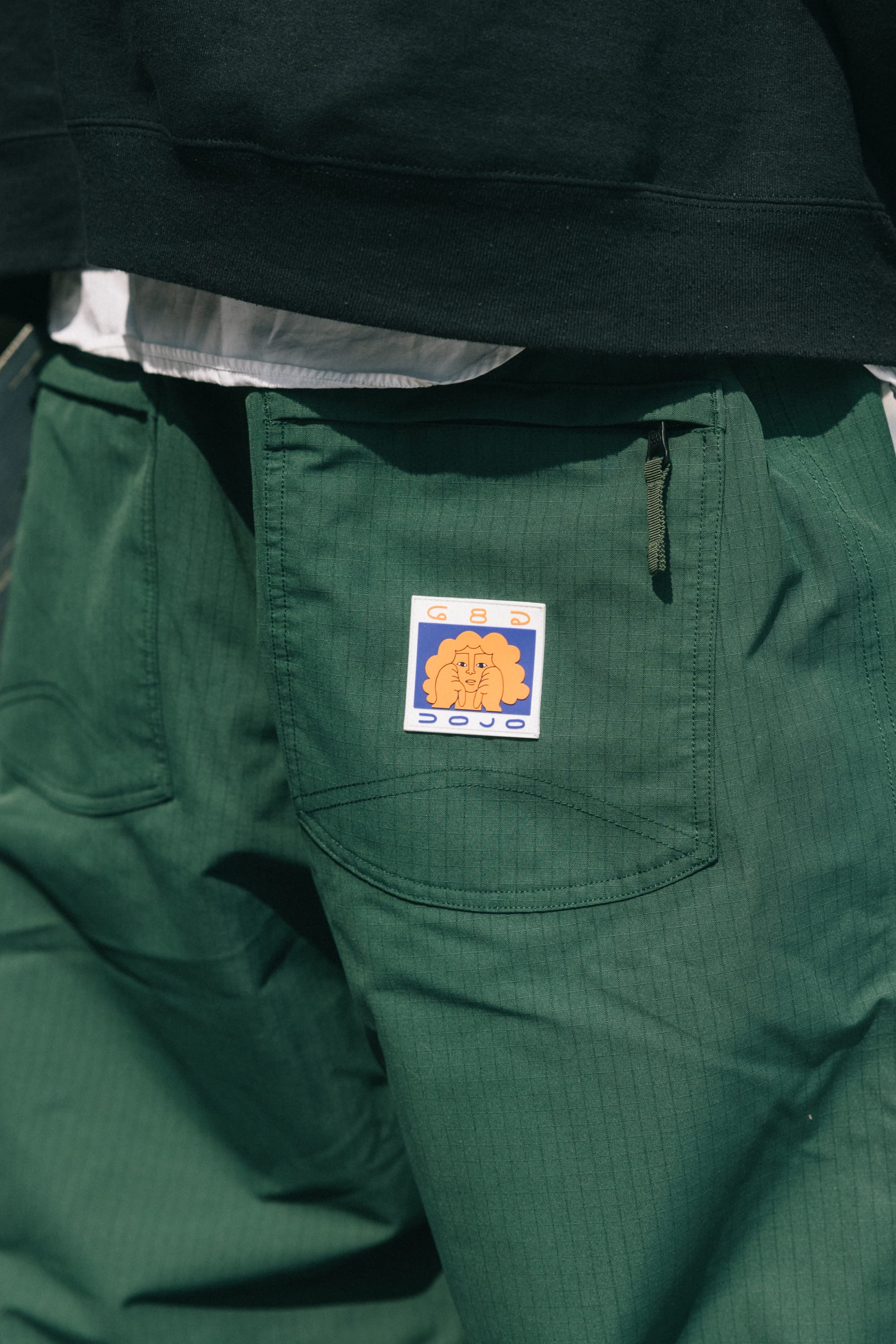 A close-up of the 686 Dojo® Pant in green shows a front pocket patch with an orange brain and stylized yellow and blue text, paired with a black shirt partially covering the waist.