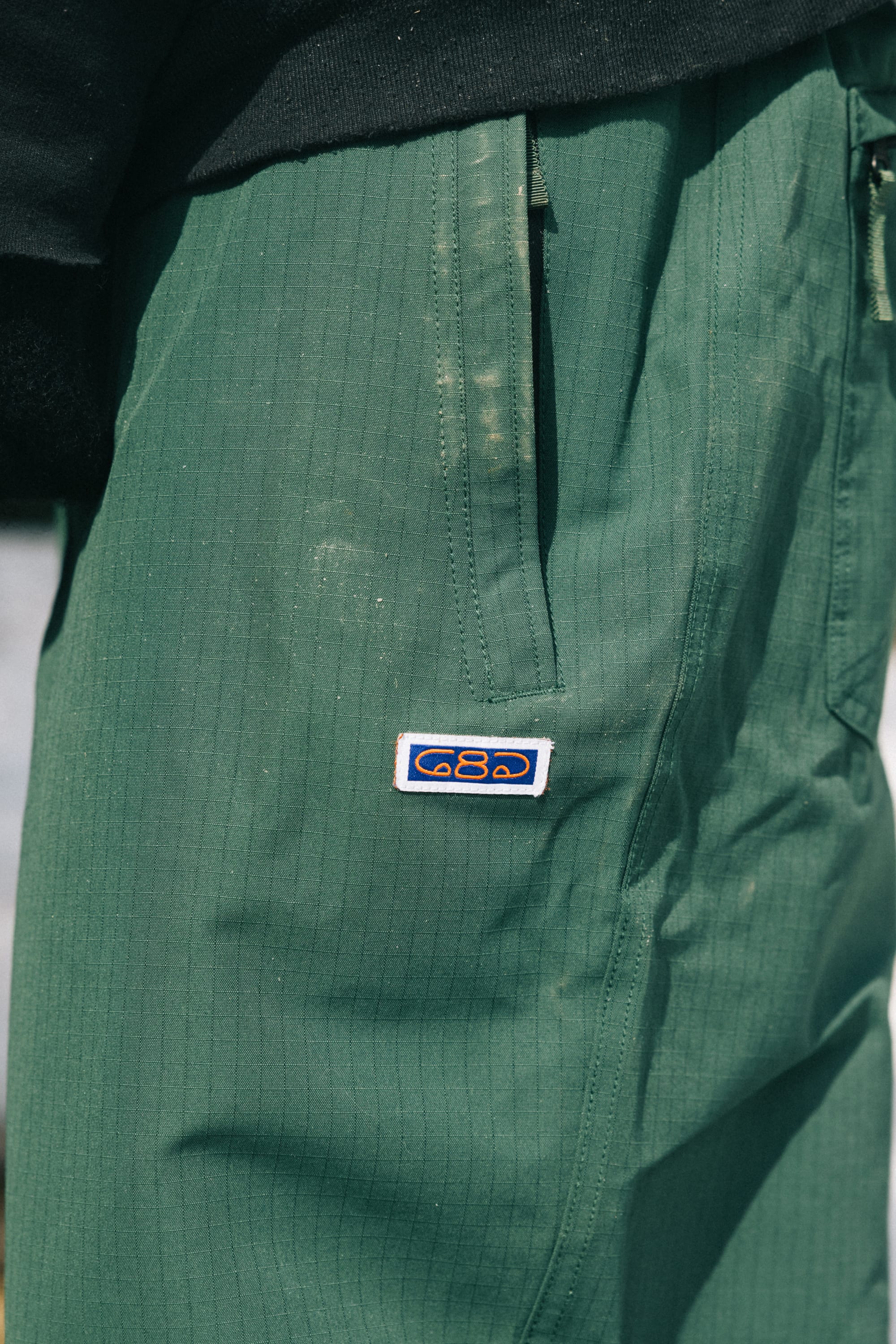Close-up of the 686 Dojo® Pant in green, showing a visible side pocket and a small rectangular patch with the blue and yellow 686 logo. These men's woven pants feature a subtle vertical stripe texture for added style.