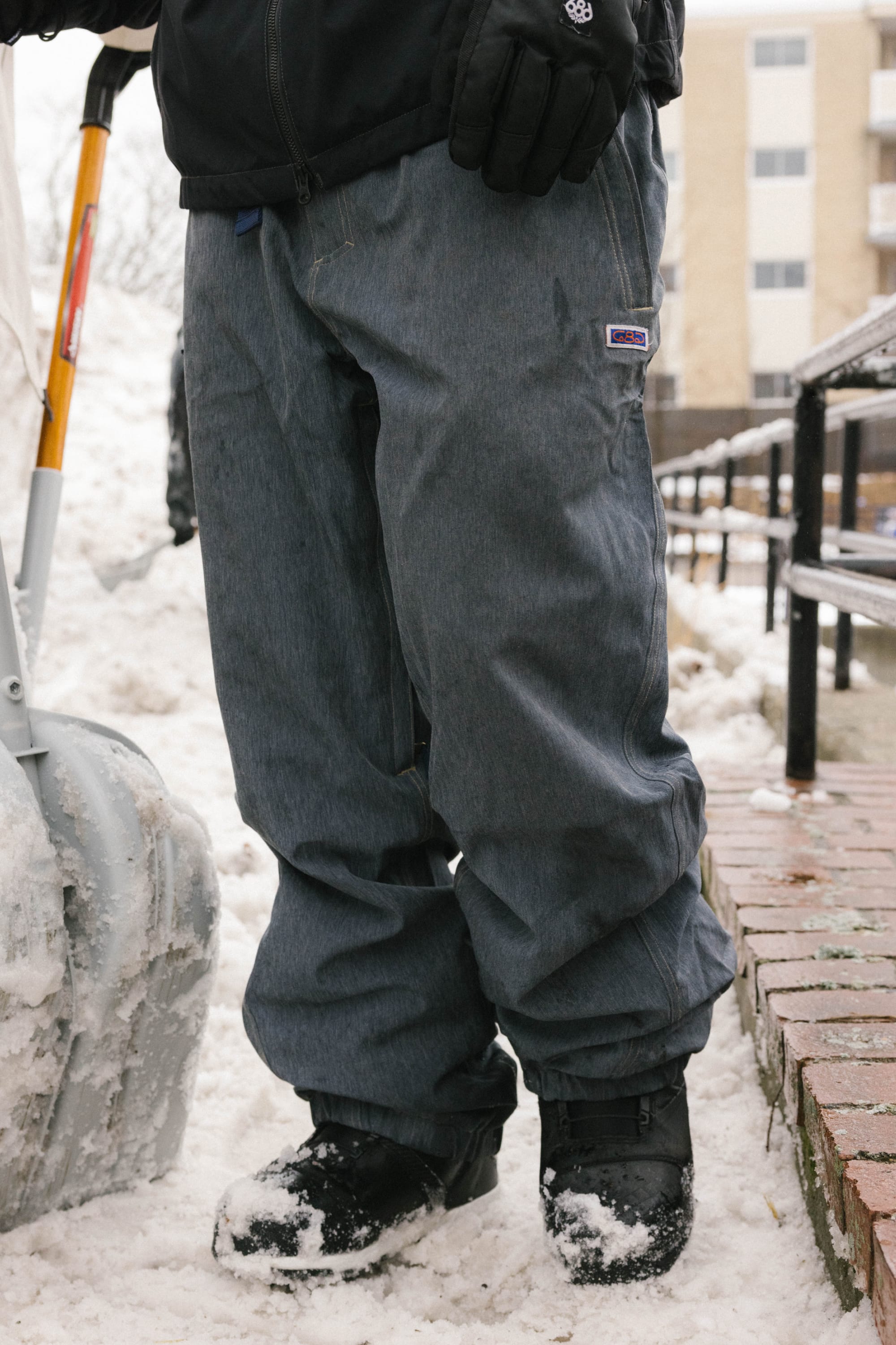 A person wearing 686 Dojo® Pant, black gloves, and boots stands in the snow with a snow shovel. Snow covers the ground and a brick ledge nearby, with buildings visible in the background.