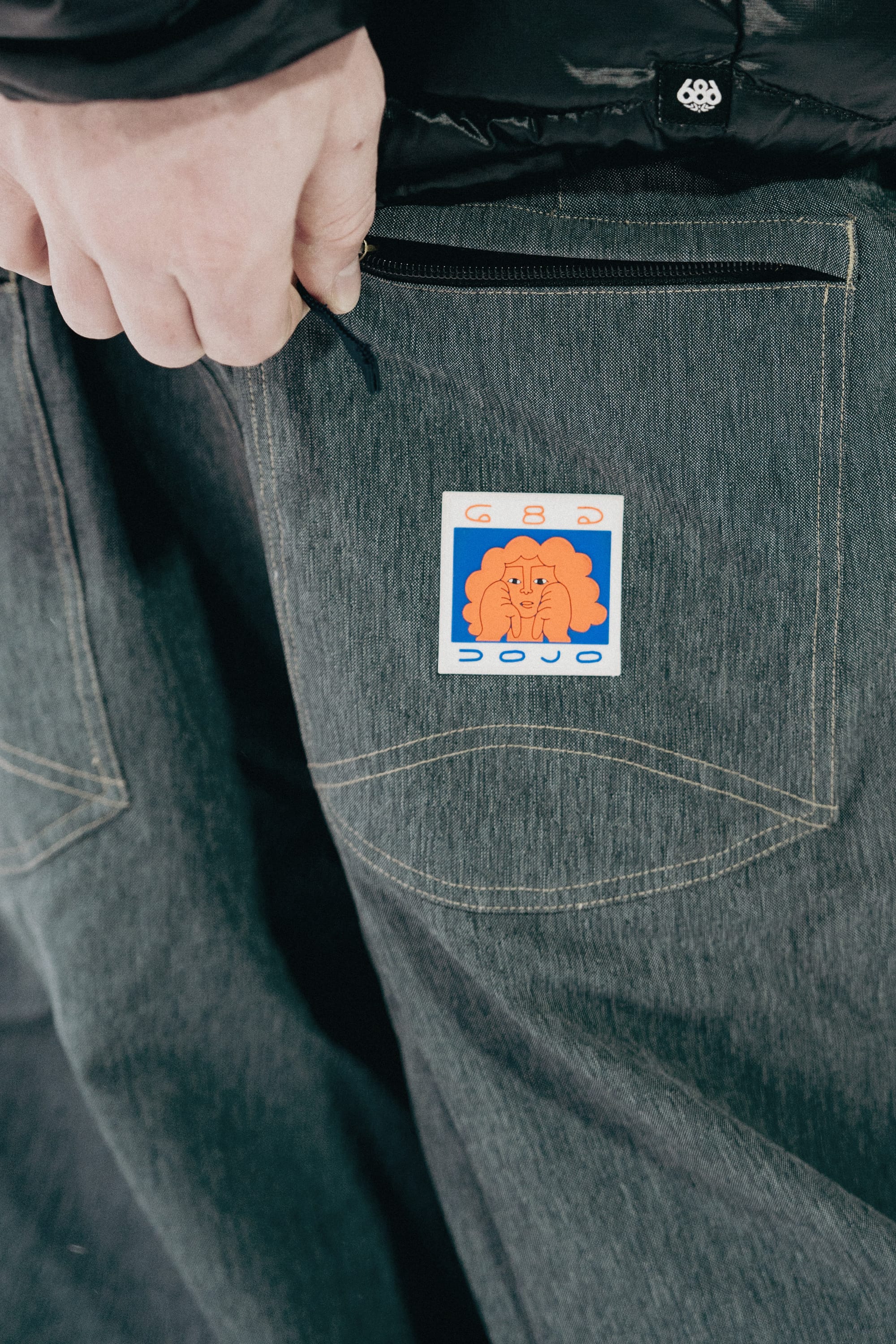 A close-up shows a hand near the back pocket of dark 686 Dojo® Pants by 686, featuring a colorful sticker with an illustrated face and the word “DOJO.”.