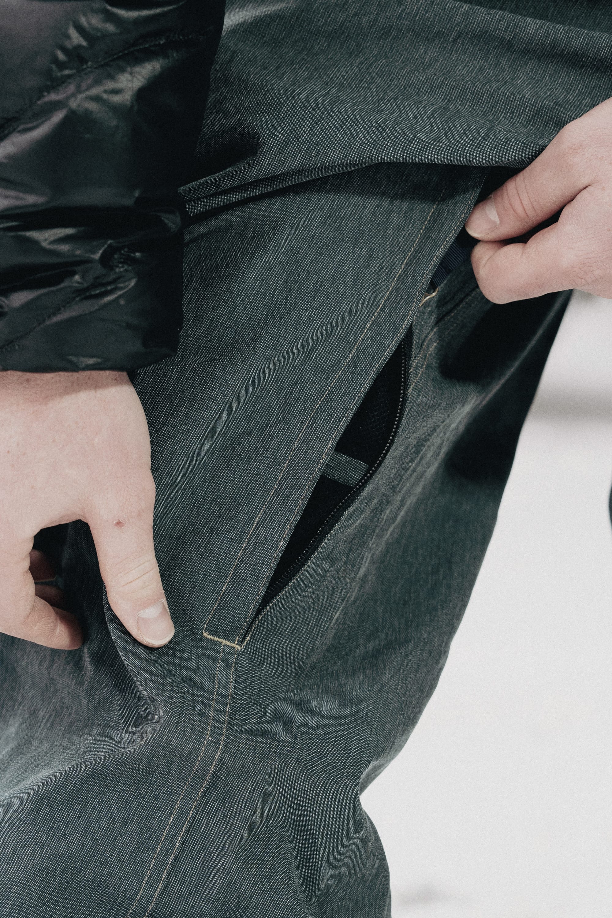 A close-up of a hand opening the zippered pocket on gray 686 Dojo® Pant men's waterproof pants, with part of a black jacket sleeve visible.