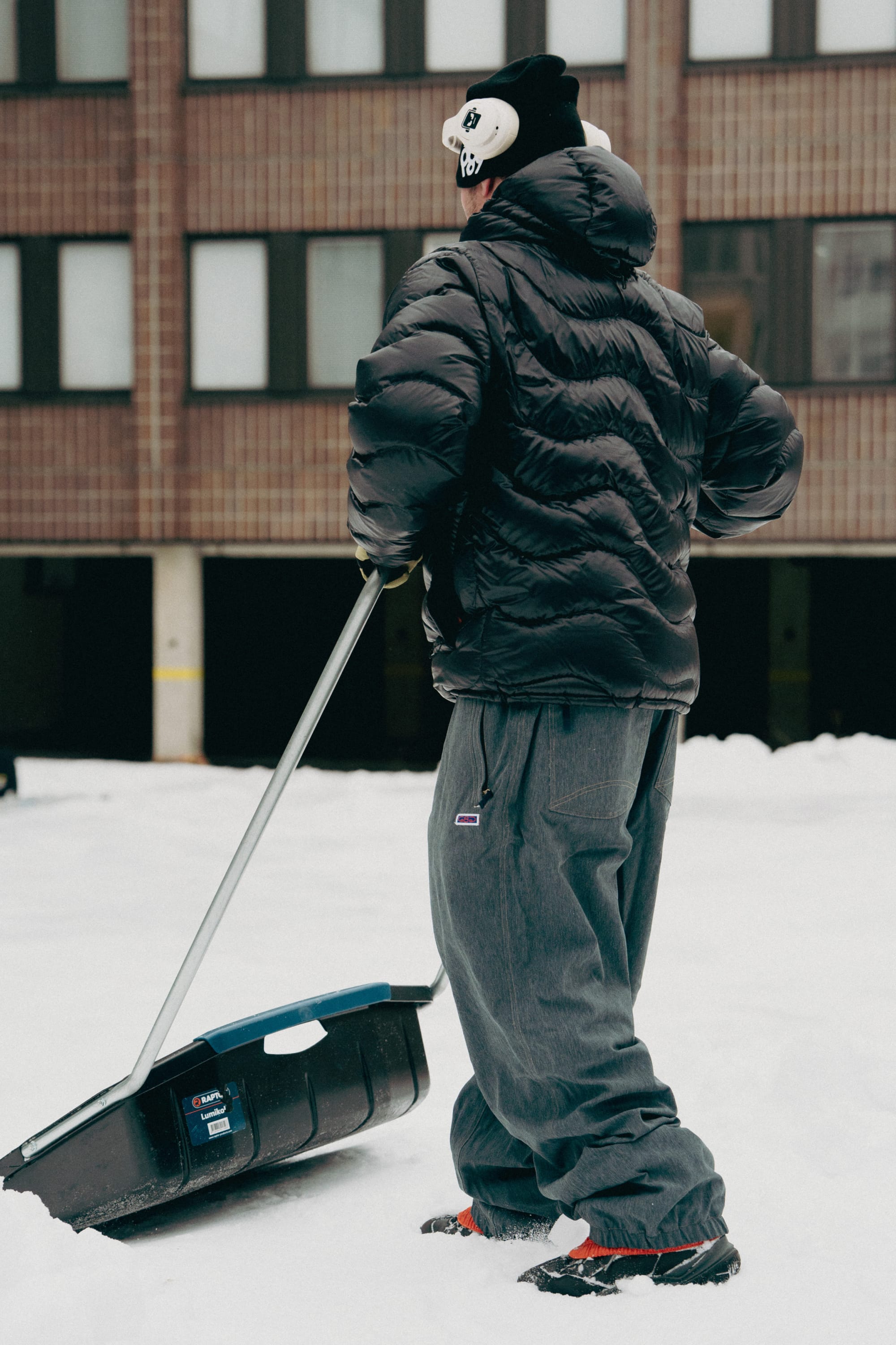 Wearing a black puffer jacket, 686 Dojo® Pant by 686, and a black beanie with a cartoon face, a person shovels snow in front of a building.