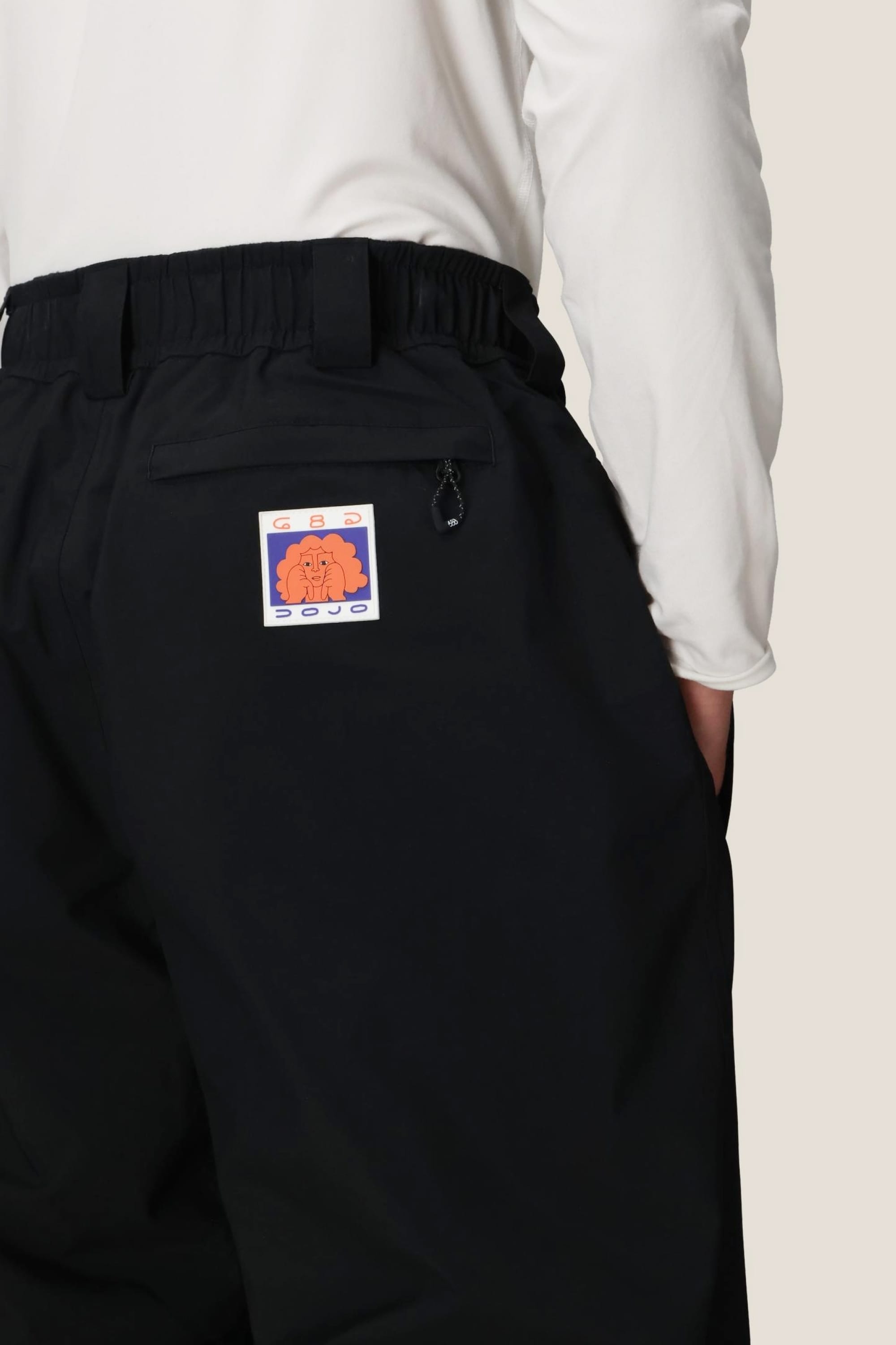A person wears the 686 Dojo® GORE-TEX Pant by 686, featuring a colorful abstract patch on the back pocket, with a white long-sleeve shirt tucked in and their hand in the pocket.