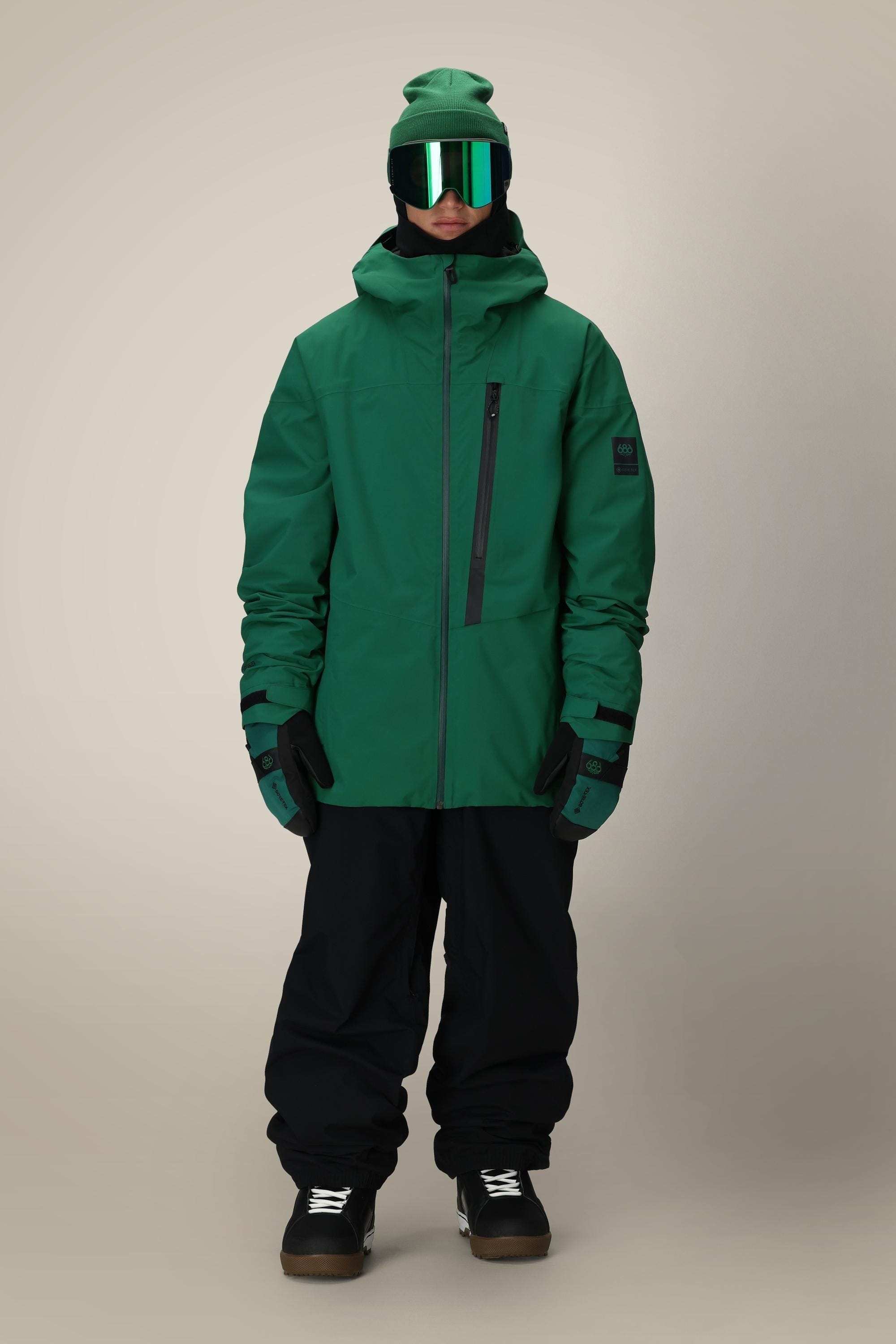 Wearing a green hooded winter jacket, 686 Dojo® GORE-TEX Pant by 686, black gloves, dark boots, a green beanie, and reflective green ski goggles, a person stands facing forward against a plain light background.