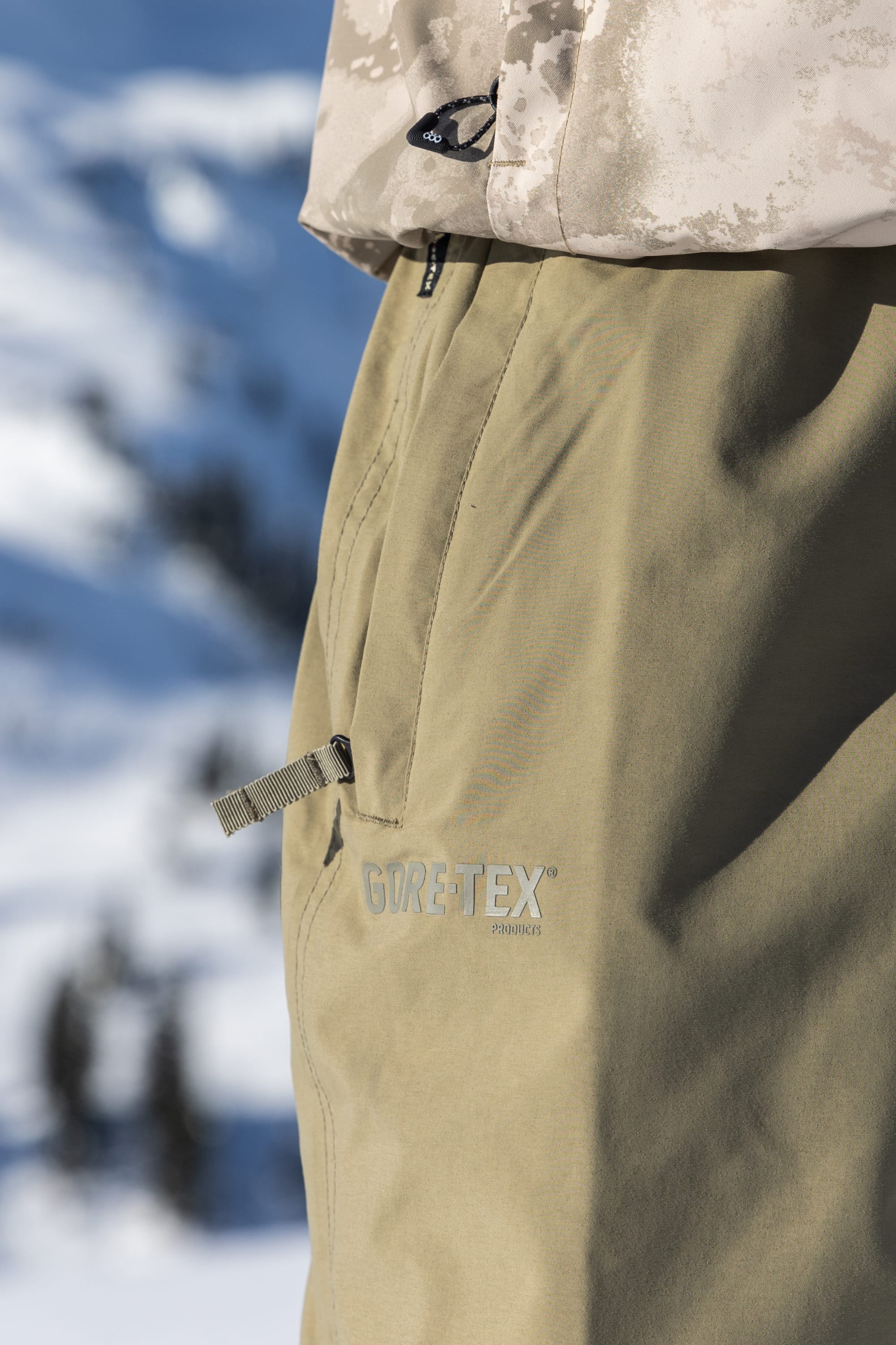 Close-up of a person wearing 686 Dojo® GORE-TEX Pant in light brown, paired with a beige patterned jacket, with snowy mountains blurred in the background.