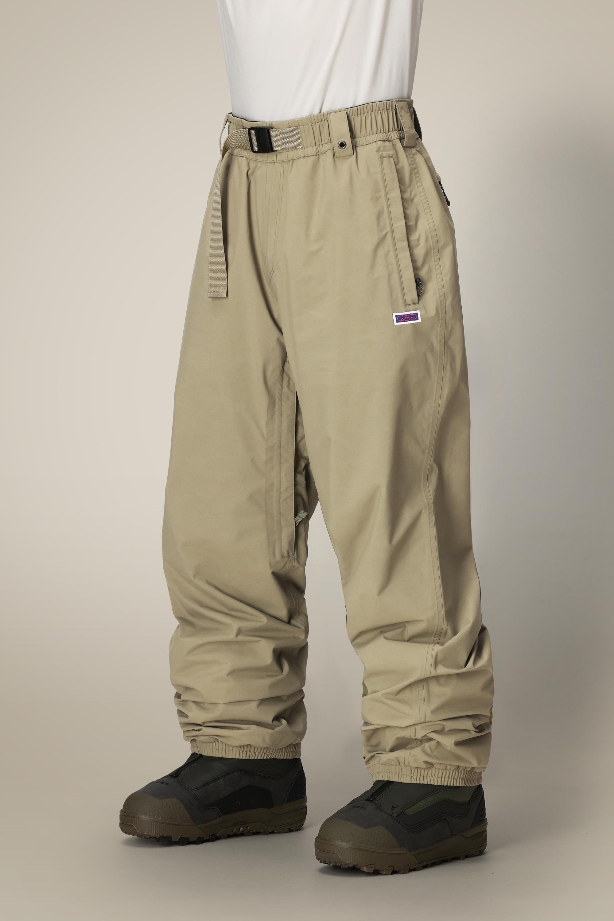 A person wearing 686 Dojo® GORE-TEX Pant by 686, tan and loose-fitting with a built-in belt, and black and olive green shoes stands against a plain light background.