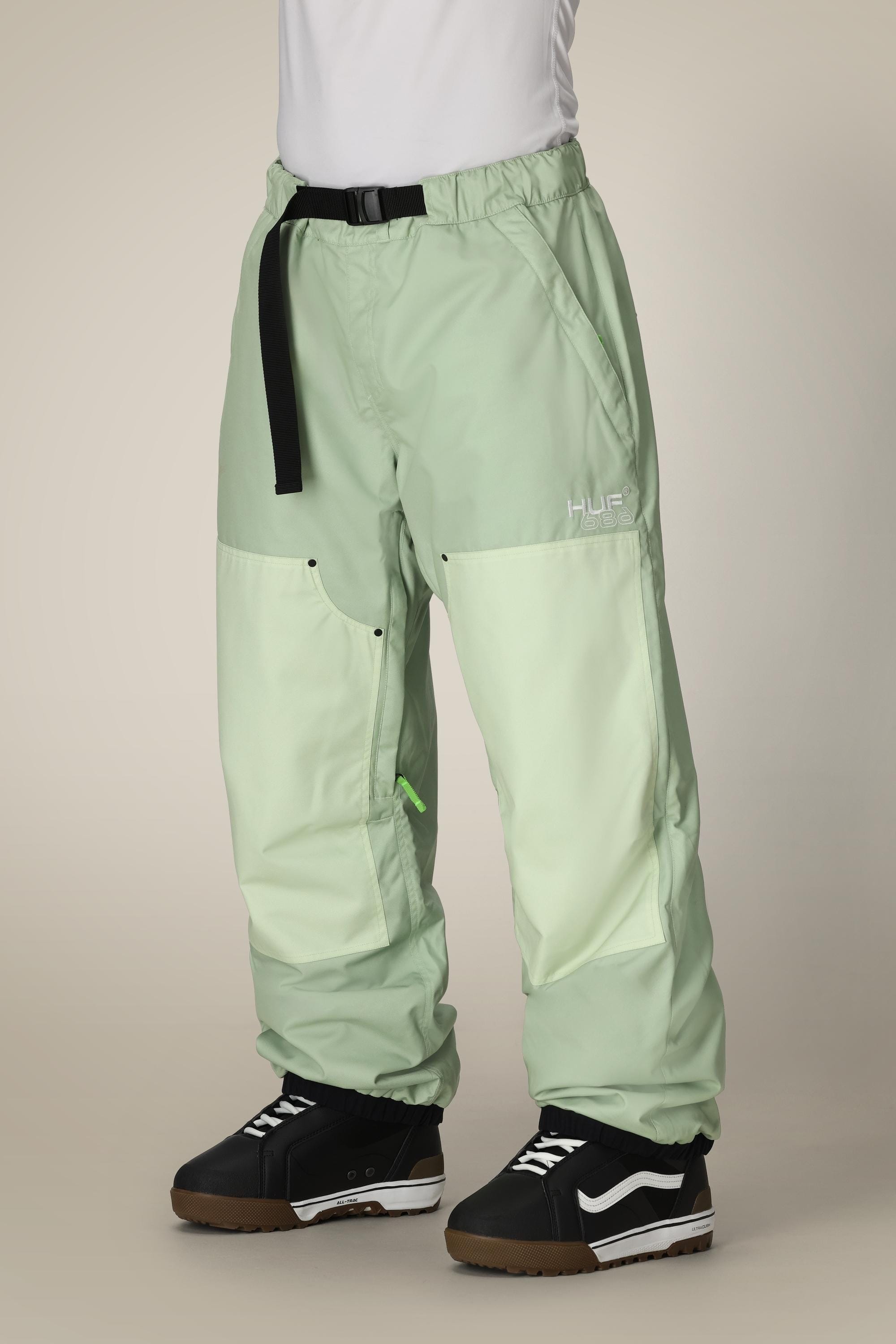 A person wears 686 Mens HUF Double H Shell Pant—loose, light green waterproof pants with a black belt and white HUF logo—paired with black and white skate shoes. The upper body is out of frame.