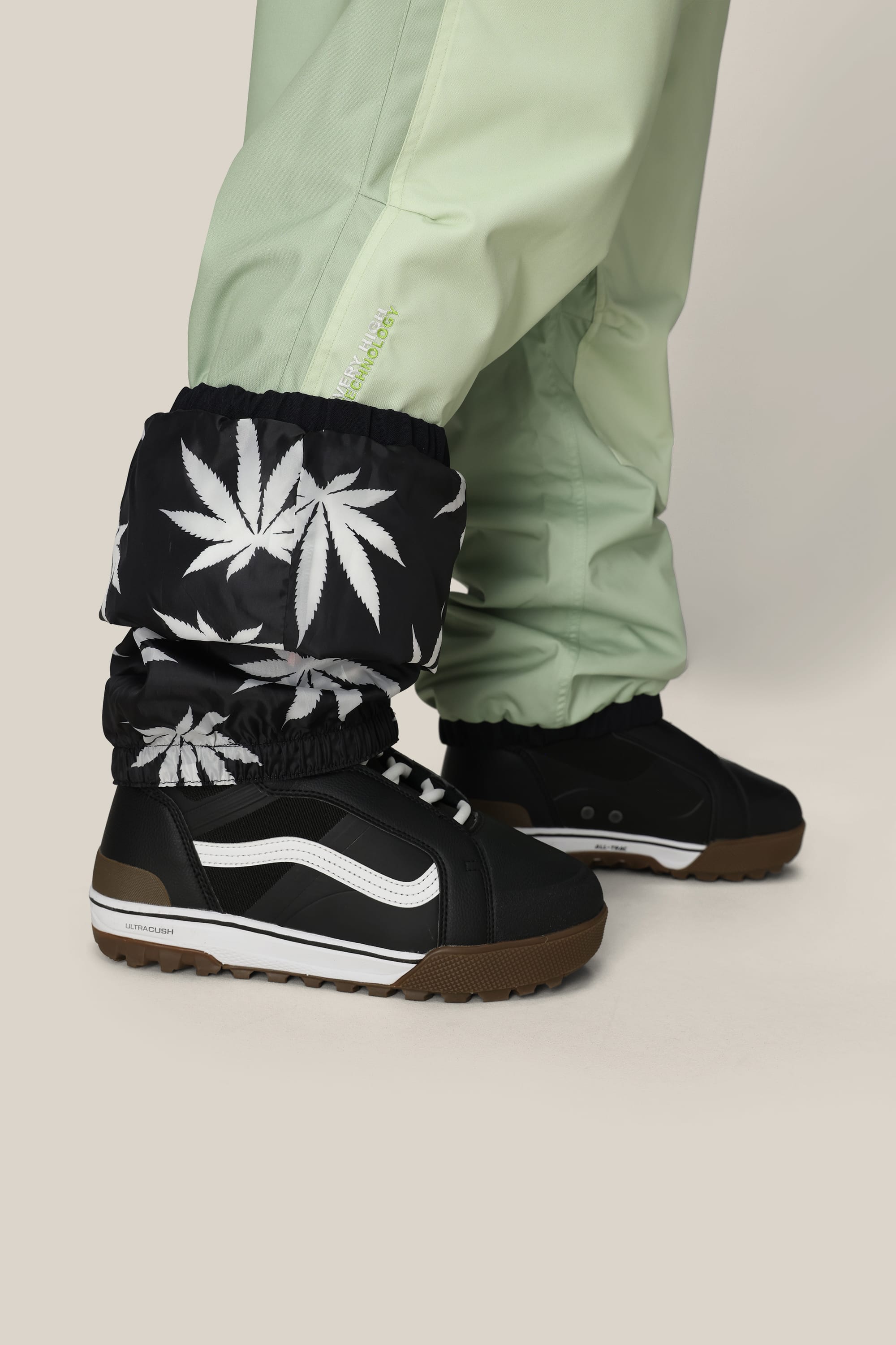 A person wearing 686 Mens HUF Double H Shell Pant in light green with black gaiters featuring white marijuana leaf prints, paired with black sneakers with white stripes and brown soles, stands against a light background.