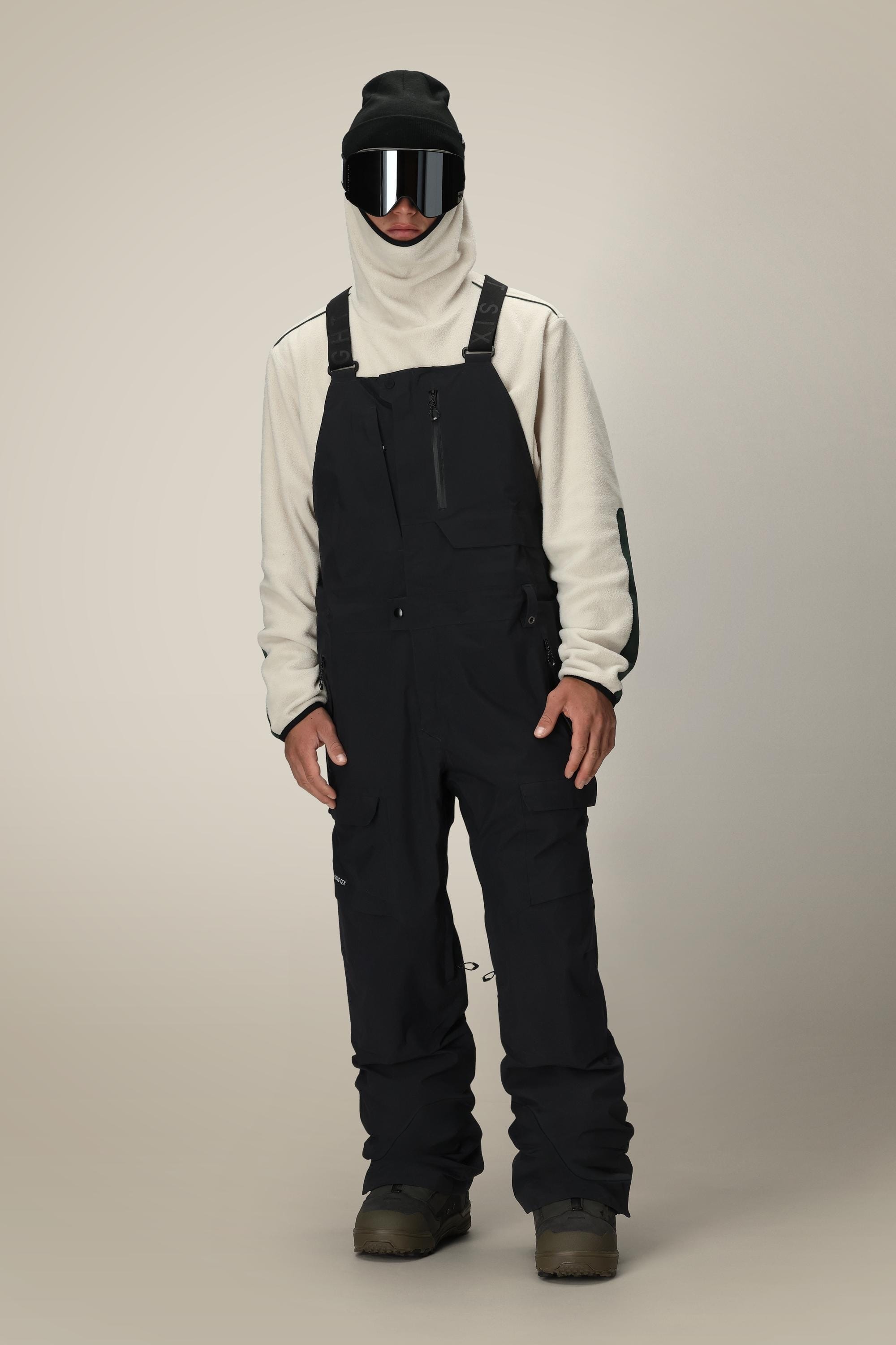 A person wearing the 686 Men’s GORE-TEX® Dispatch™ Shell Bib, a white hoodie, reflective ski goggles, a black beanie, and brown boots stands against a plain beige background.