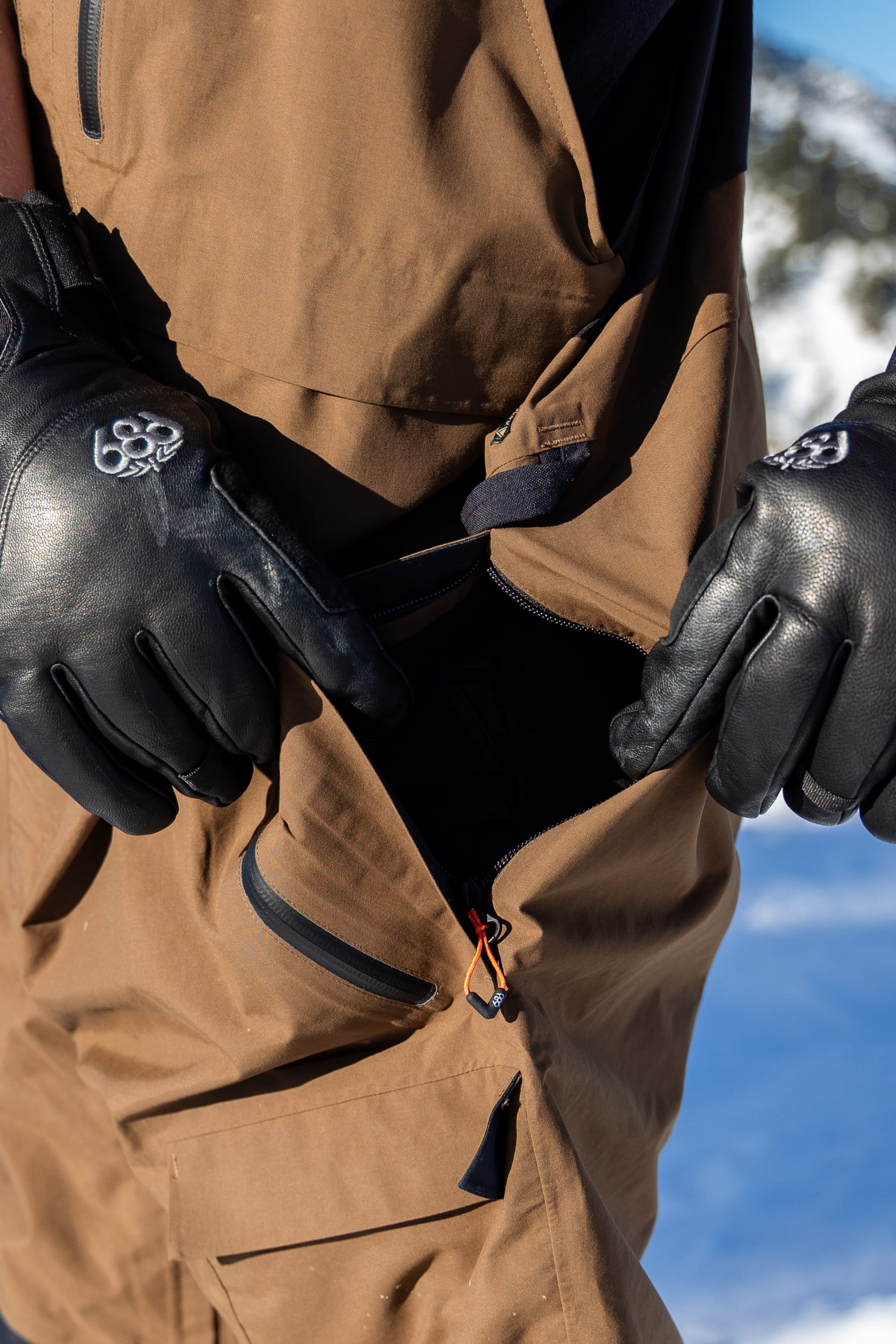 A person wearing black gloves unzips the 686 Men’s GORE-TEX® Dispatch™ Shell Bib, with snow and mountains visible in the blurred background.