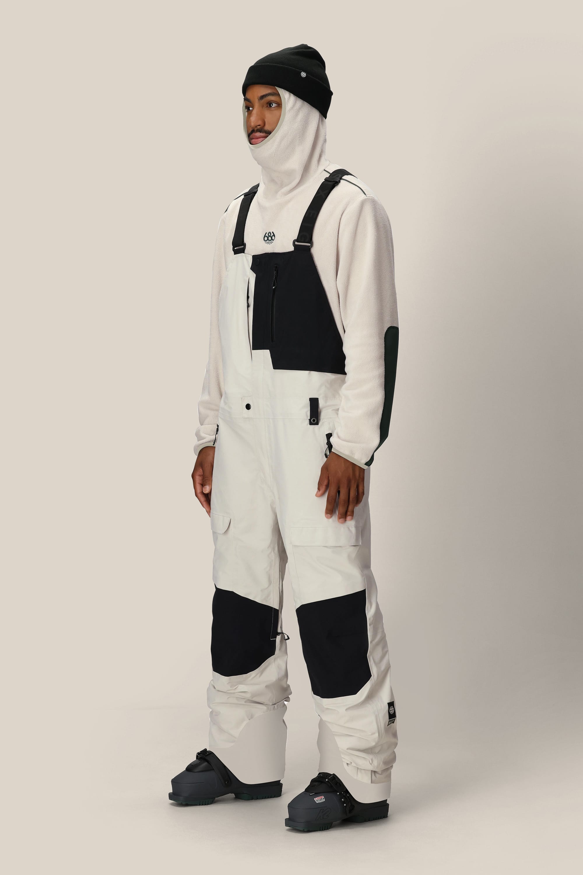 A person wearing a beanie, balaclava, snow boots, and the 686 Men's GORE-TEX Dispatch™ Shell Bib from 686 stands against a plain light background.