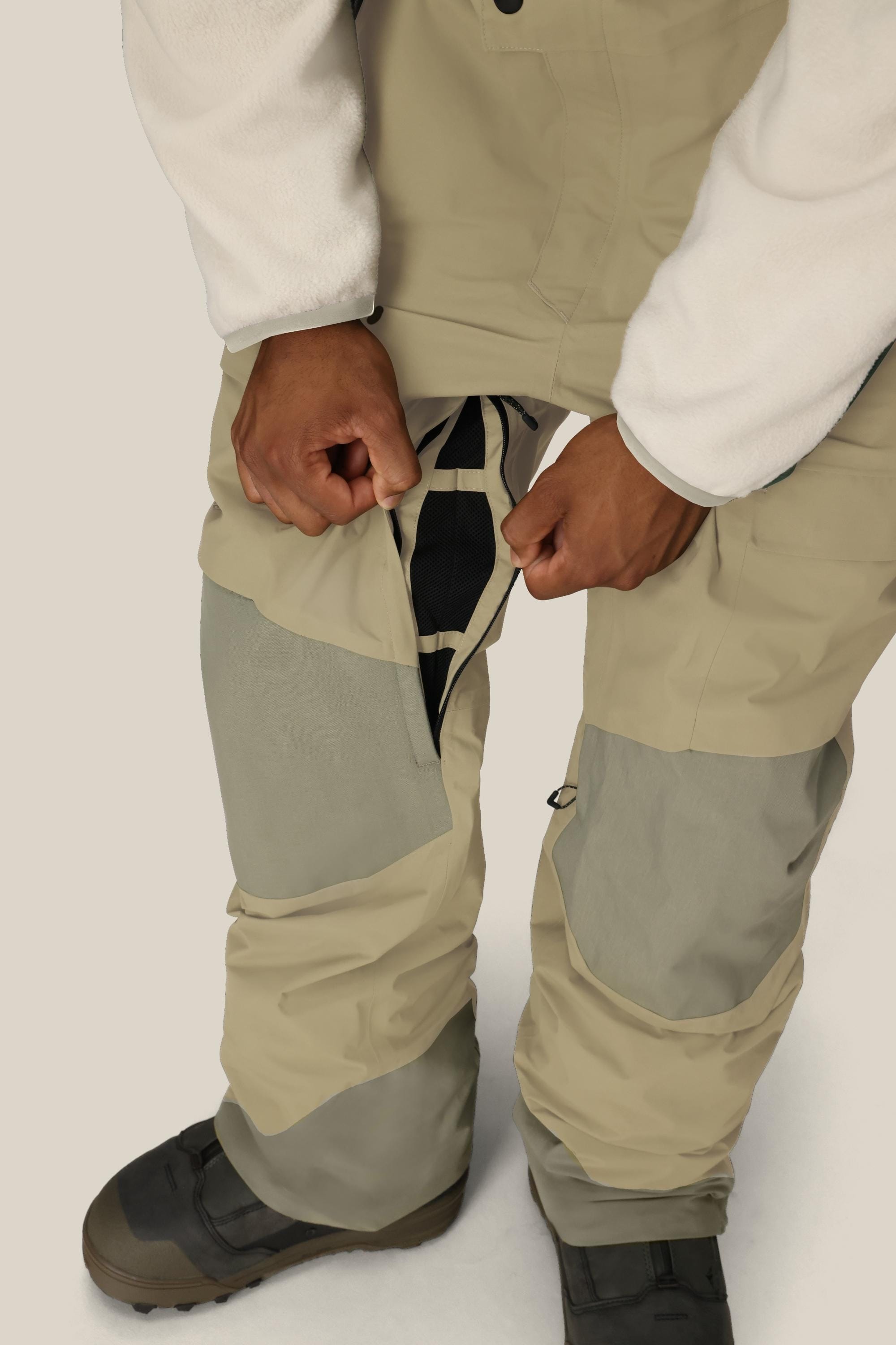 Wearing the 686 Men's GORE-TEX Dispatch™ Shell Bib in beige and gray, a person opens a mesh-lined zipper vent on the thigh, paired with a white fleece top and black boots.