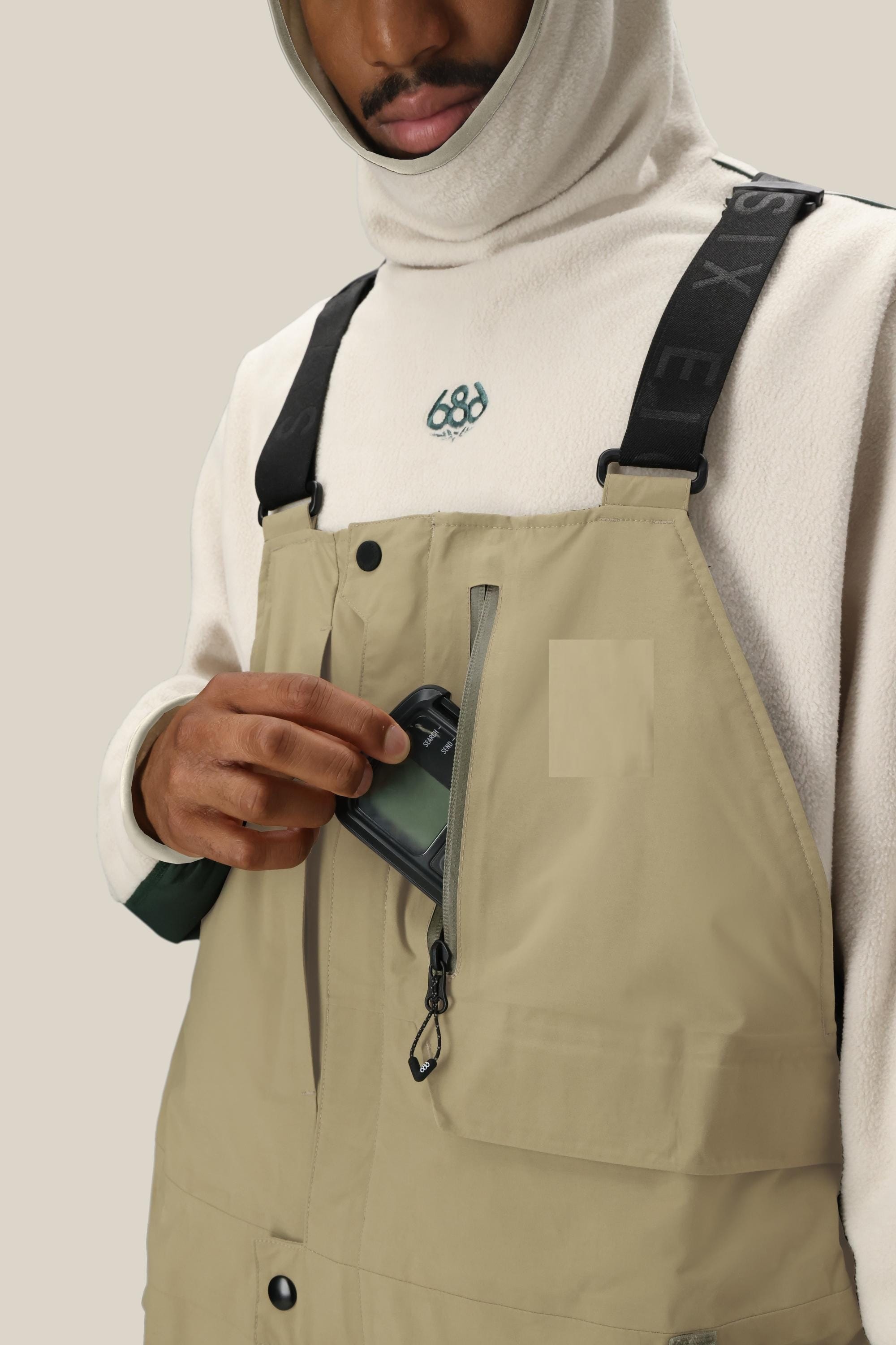 A person in a light hoodie and tan 686 Men's GORE-TEX Dispatch™ Shell Bib with black straps places a smartphone into the zippered chest pocket. Only their torso and lower face are visible.