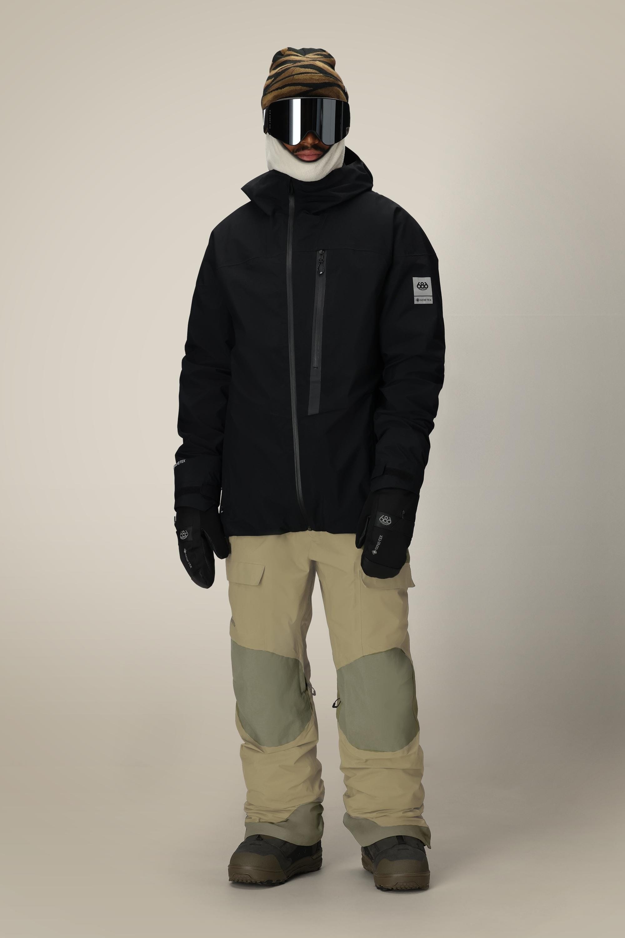 A person stands against a plain light background wearing the 686 Men's GORE-TEX Dispatch™ Shell Bib from 686, paired with a brown striped beanie, large ski goggles, black jacket, black gloves, and snow boots.