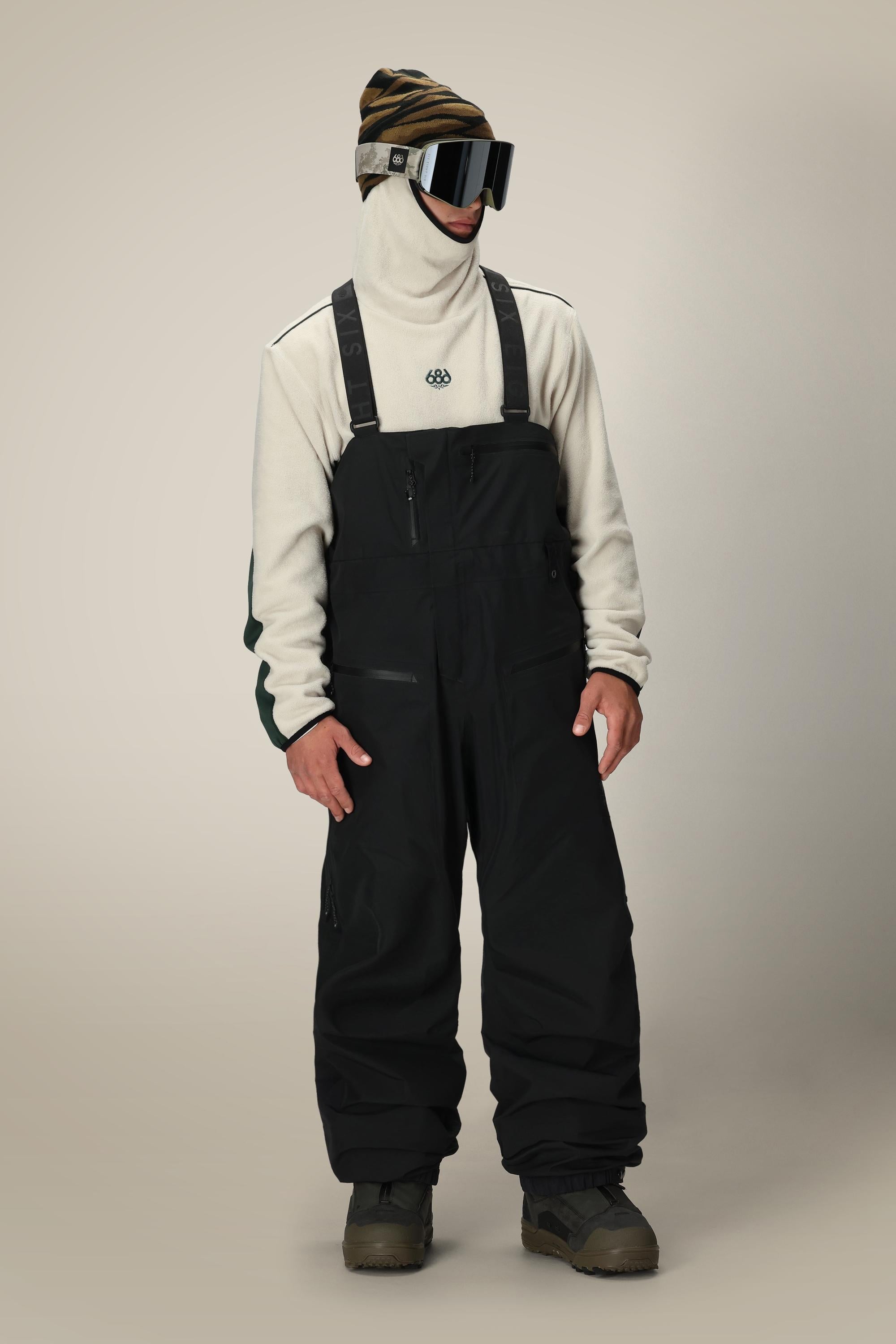 A person wearing the 686 Mens GORE-TEX 3L ATV Bib by 686, a cream high-neck pullover, beanie, large ski goggles, and dark boots stands against a plain light background.