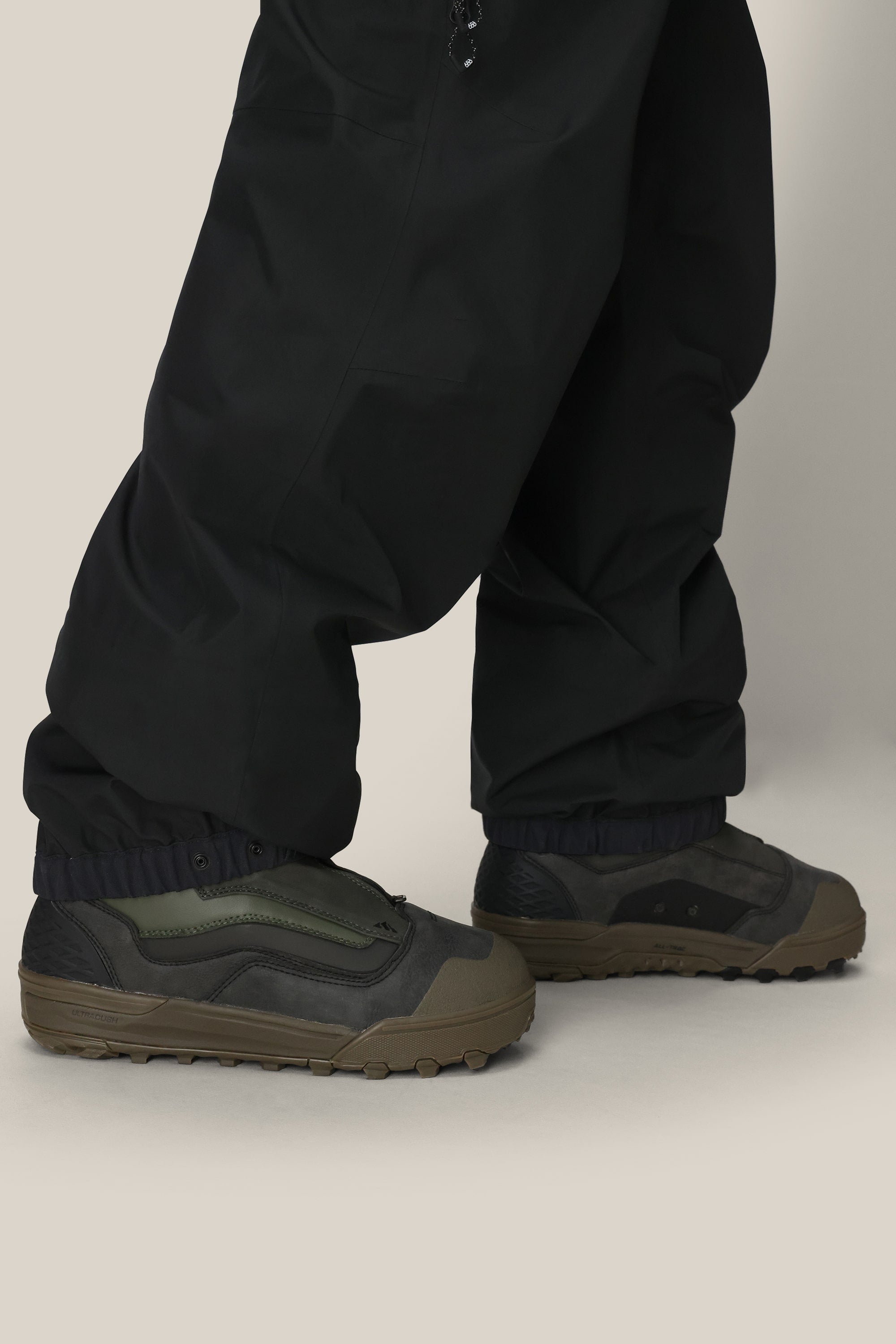 Someone stands on a light surface wearing 686 Mens GORE-TEX 3L ATV Bib pants by 686, their loose black legs draping over dark green and black sneakers, slightly covering the tops.