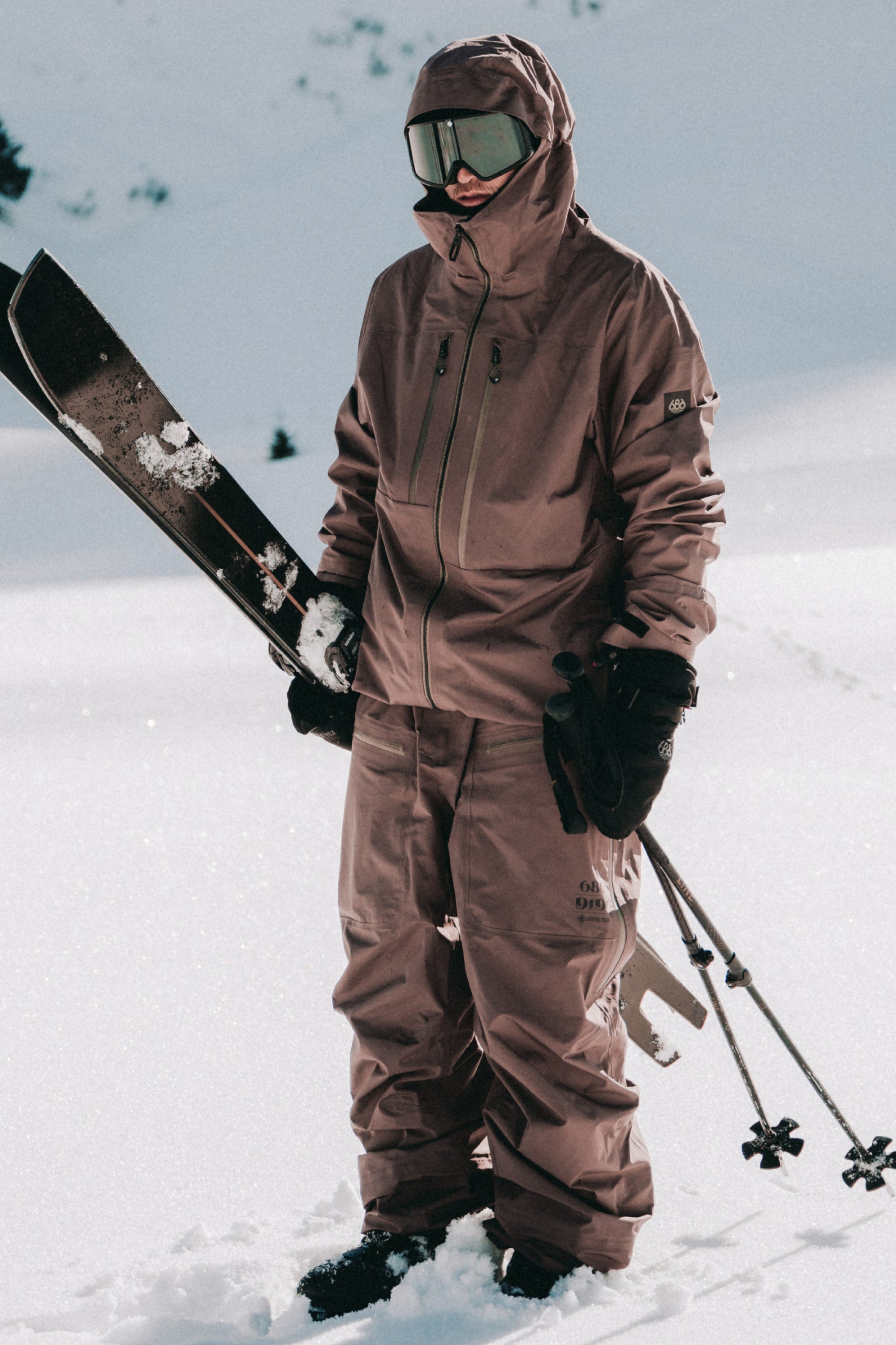 A person stands on snowy ground in 686 Mens GORE-TEX 3L ATV Bib by 686, wearing a brown ski jacket, pants, gloves, and goggles while holding skis and poles against a stunning winter mountain backdrop.