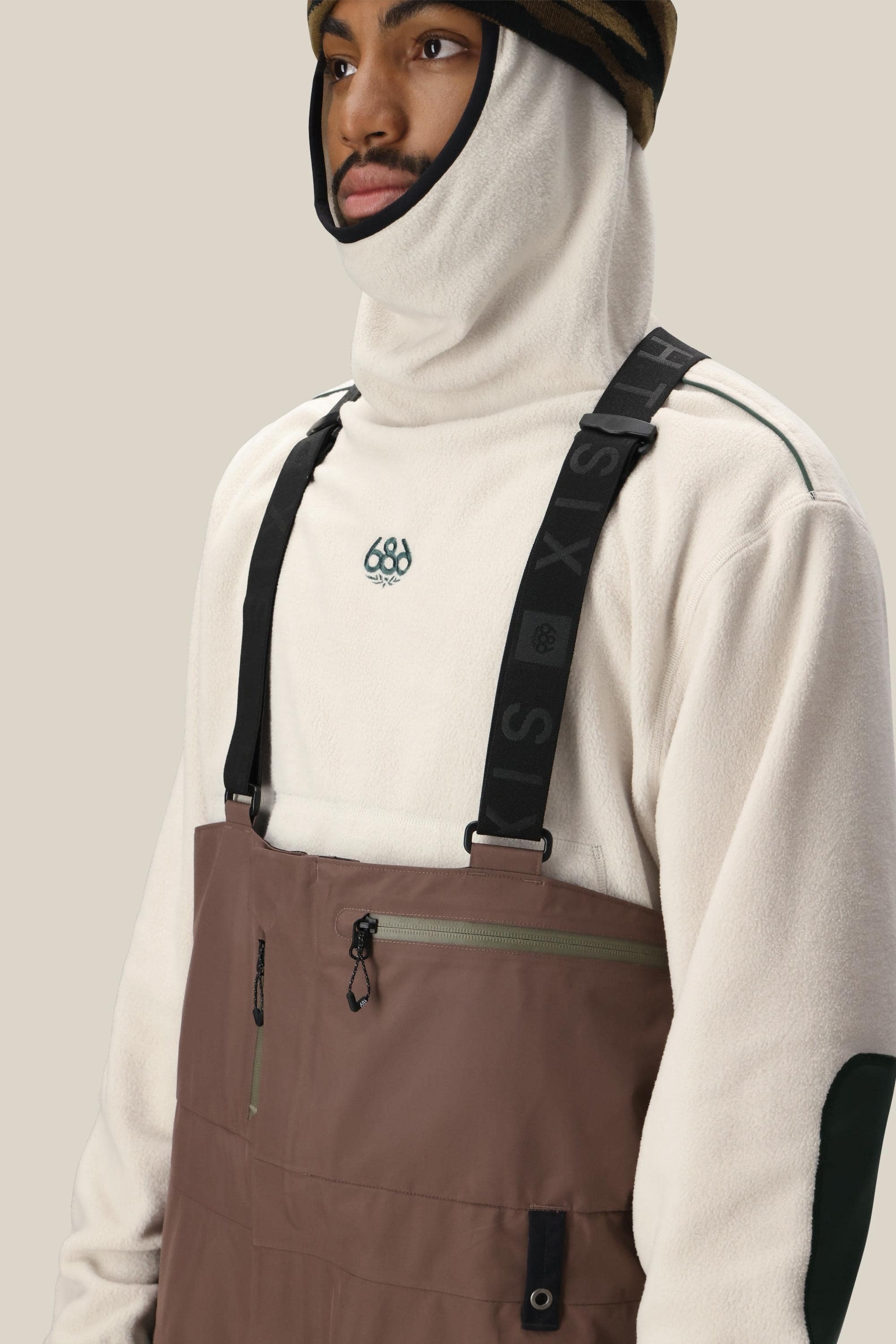 A person wearing a white fleece hoodie with a logo, the 686 Mens GORE-TEX 3L ATV Bib brown overalls with black suspenders, and a camouflage beanie, looking off to the side against a neutral background.