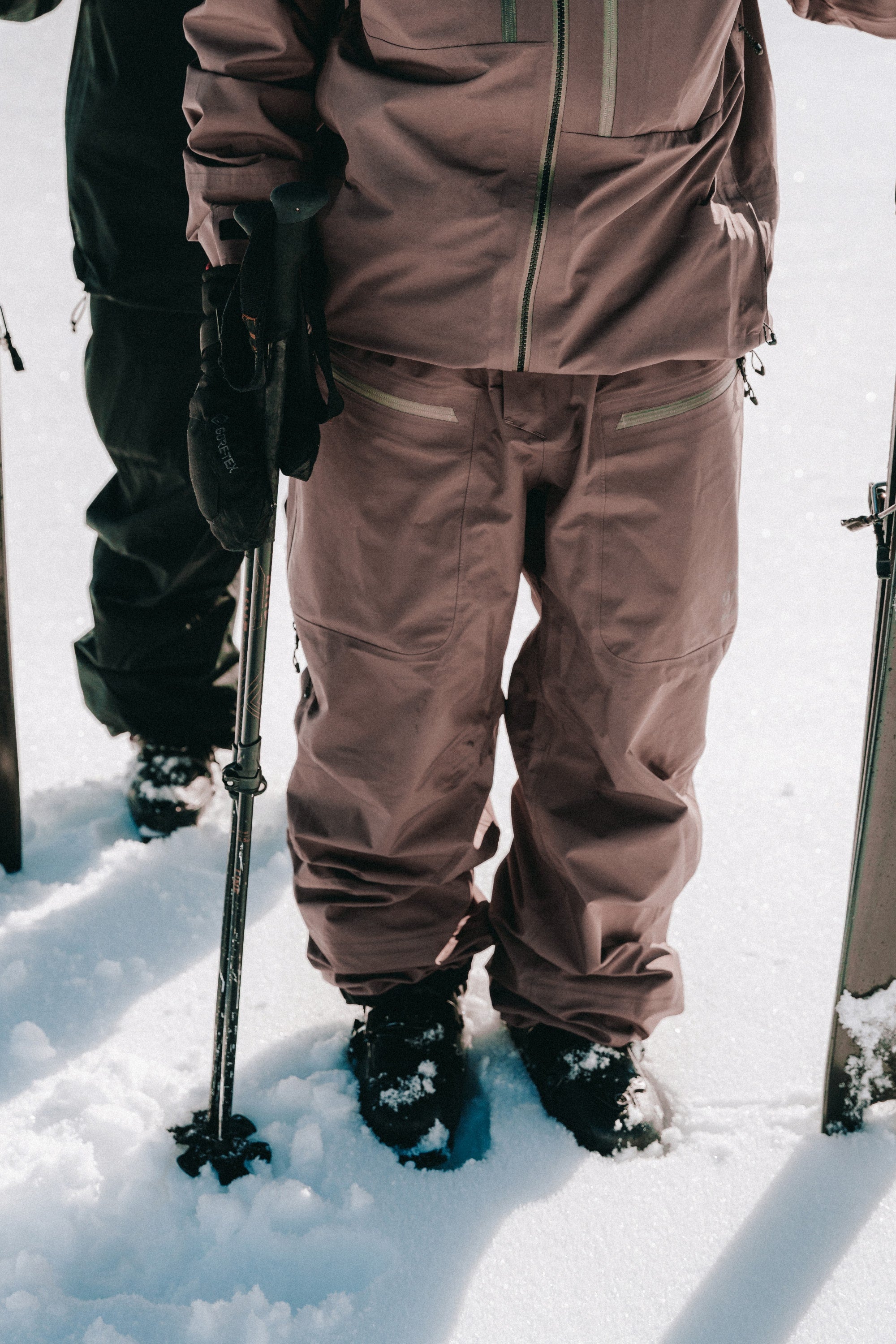 Two people stand in the snow, their lower bodies visible as they wear 686 Mens GORE-TEX 3L ATV Bibs and winter jackets, holding ski poles next to their skis.