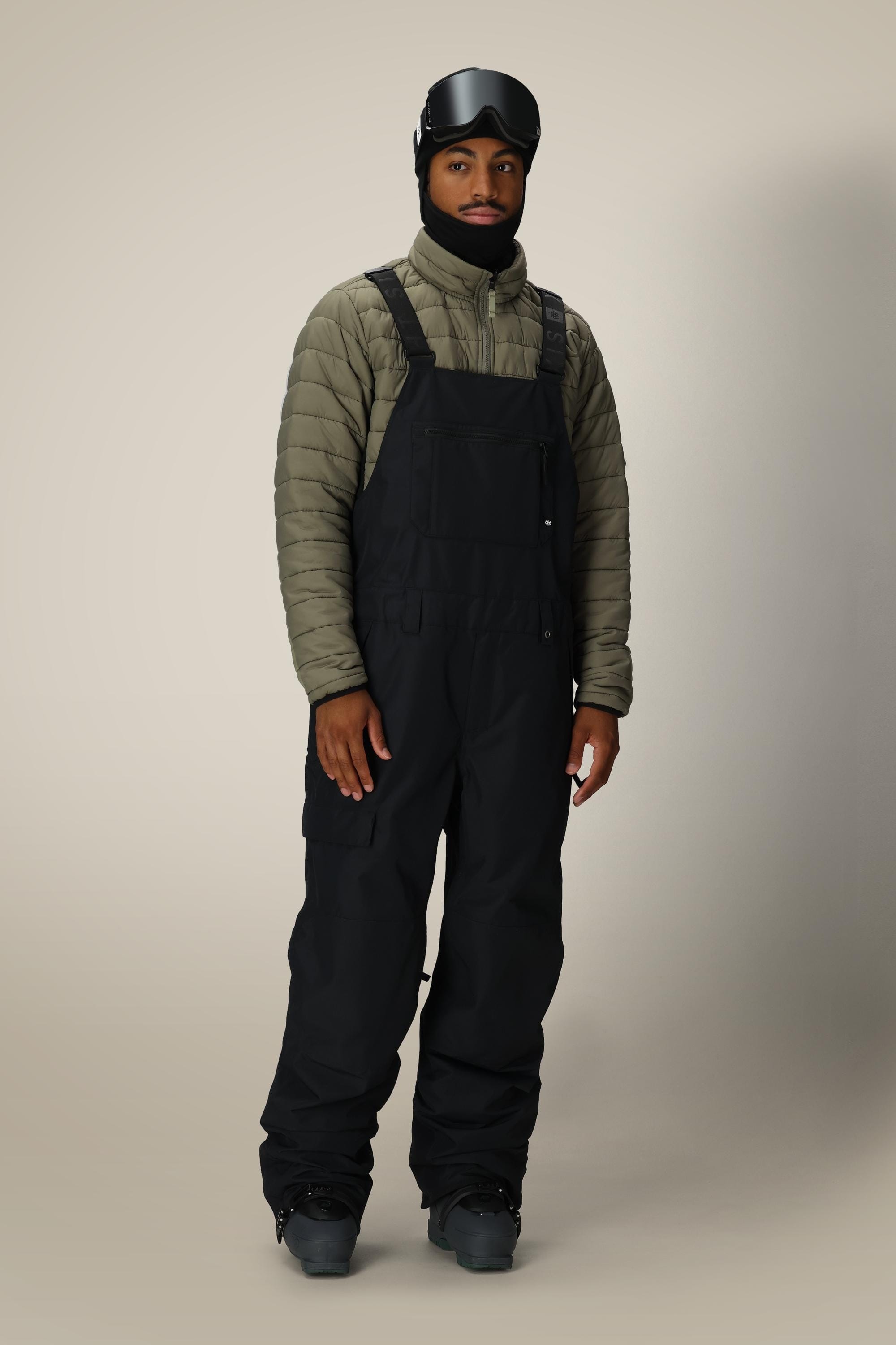 A person wearing a green puffer jacket, 686 Men's 20K Hot Lap™ Shell Bib by 686, black winter boots, a black helmet, and ski goggles stands against a plain light background.