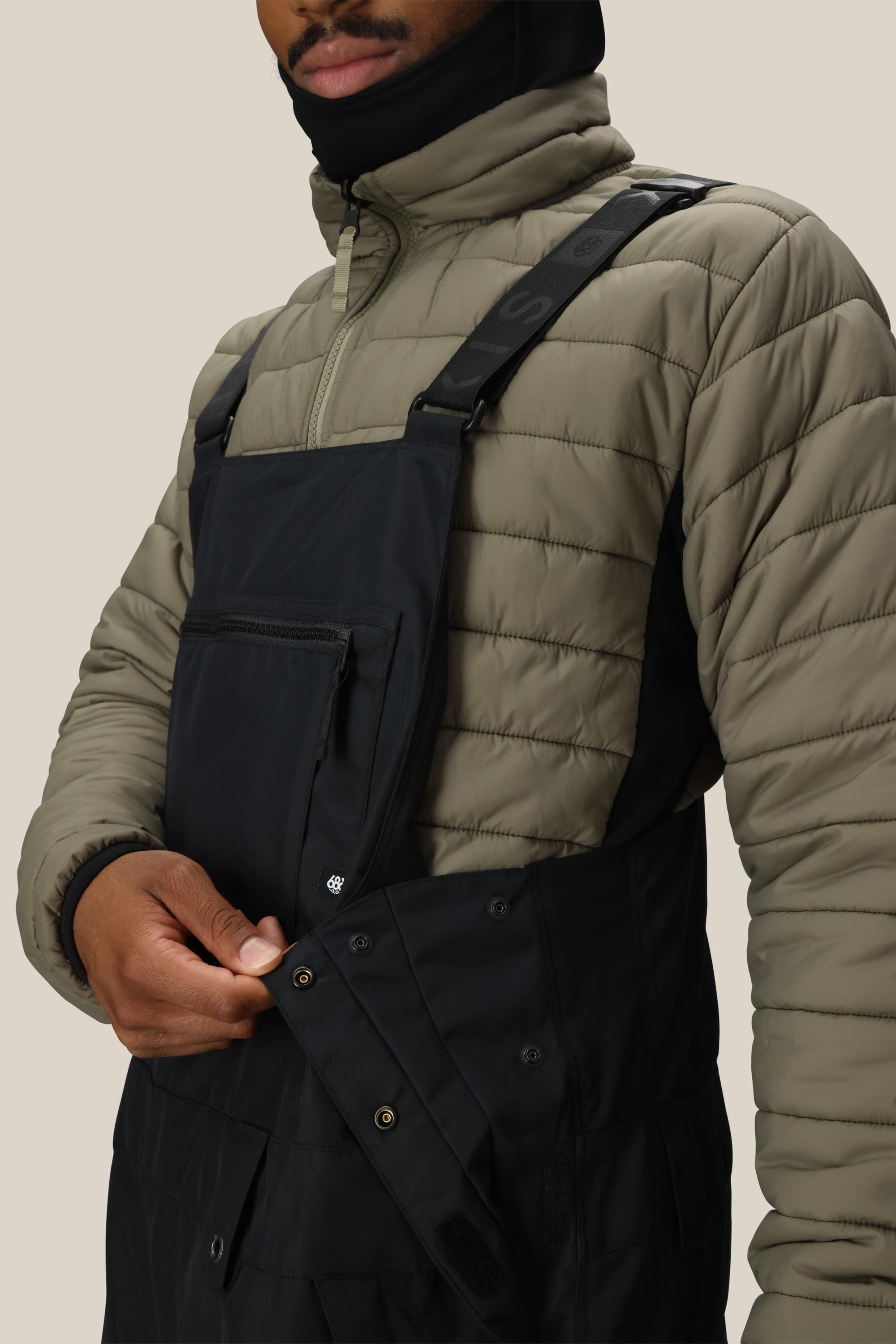 Someone wearing a light olive quilted jacket and 686 Men's 20K Hot Lap™ Shell Bib black overalls by 686 unfastens a side button on the bib; background is plain beige.