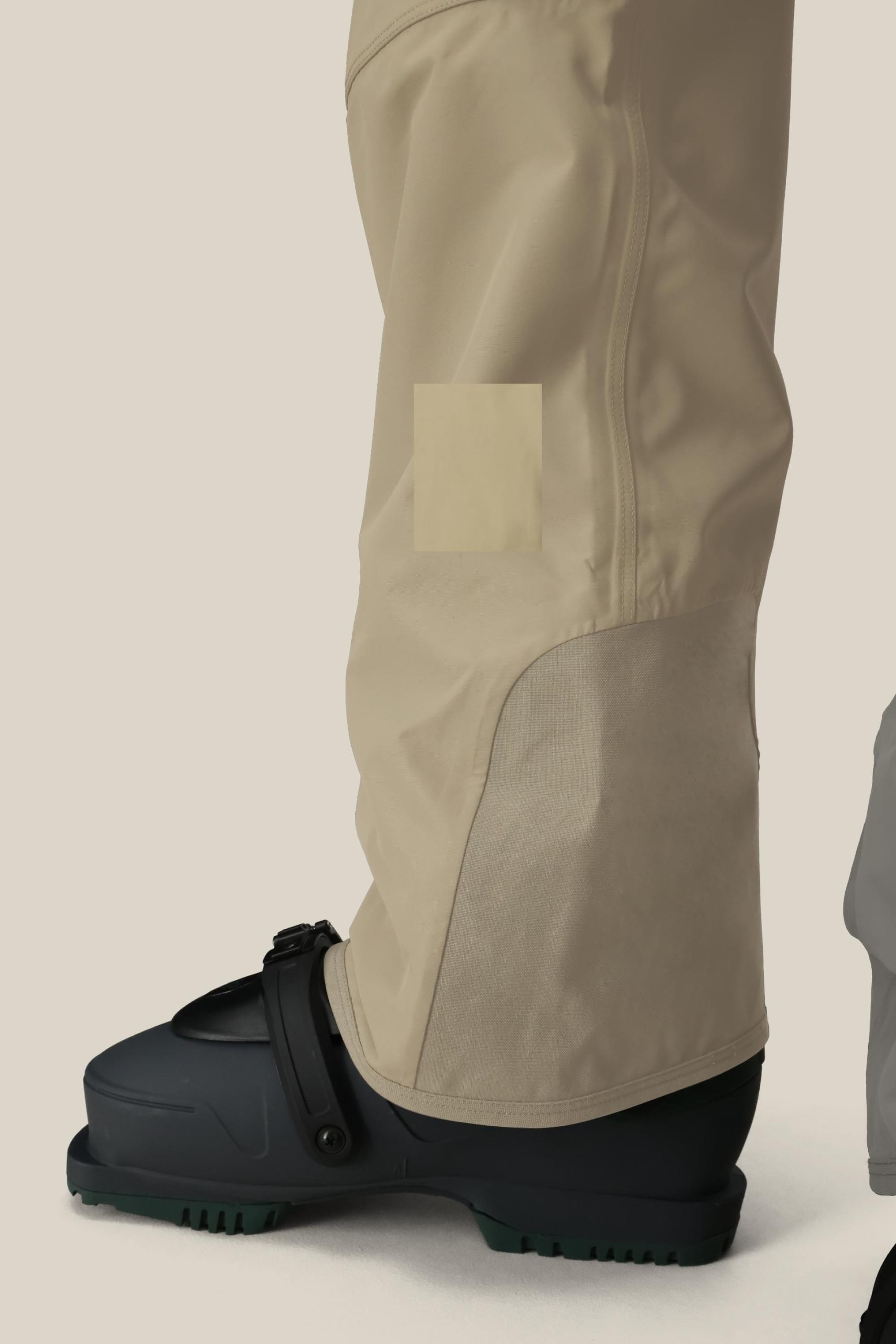 A close-up shows the lower leg and foot of a person wearing 686 Men’s 20K Hot Lap™ Shell Bib pants and a black ski boot with buckle, highlighting the detailed stitching on the beige 686 bib against a light background.