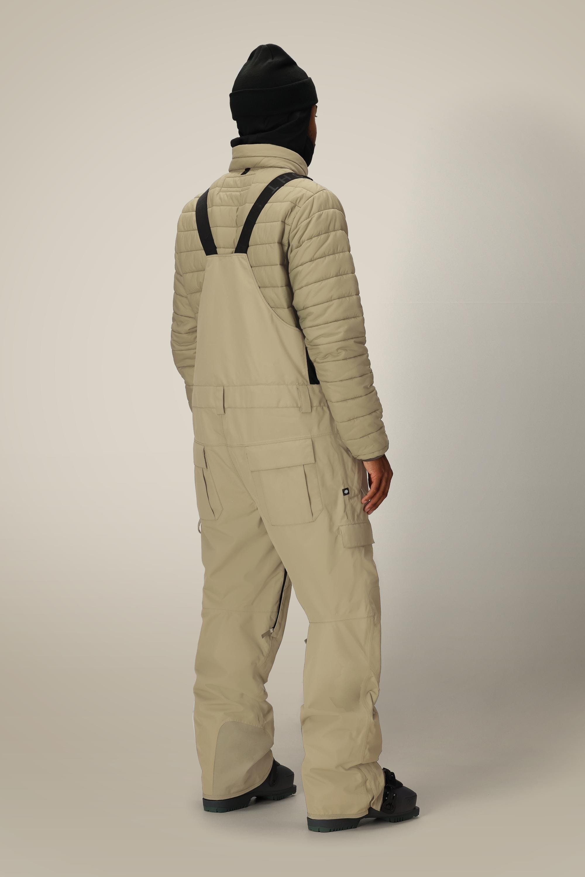 A person wearing a black beanie, a light quilted jacket, and 686 Men's 20K Hot Lap™ Shell Bib from 686 with black straps stands facing away from the camera against a neutral background.